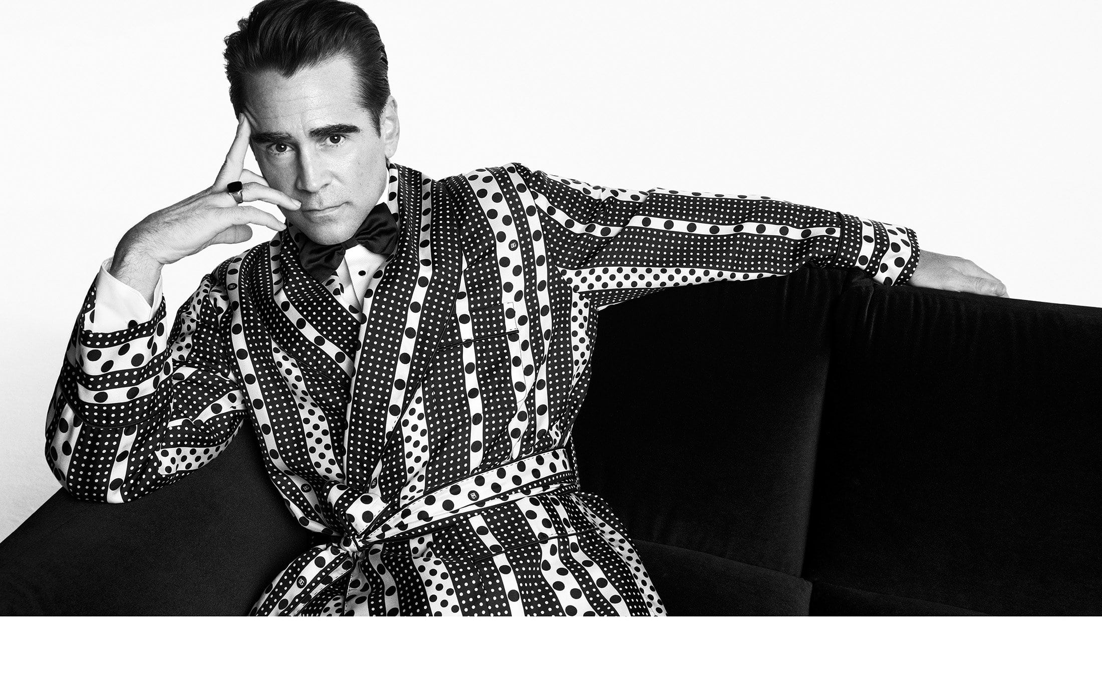 Black and white portrait of a man in a polka dot robe sitting on a black sofa, looking at the camera with a serious expression.