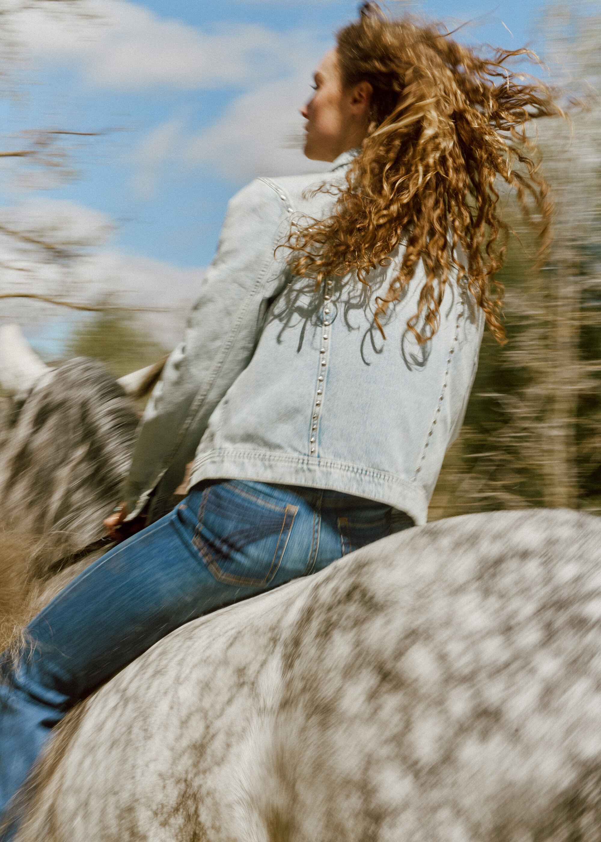 A woman with curly brown hair rides a gray dapple horse in motion through a wooded area under a bright blue sky with wispy clouds.