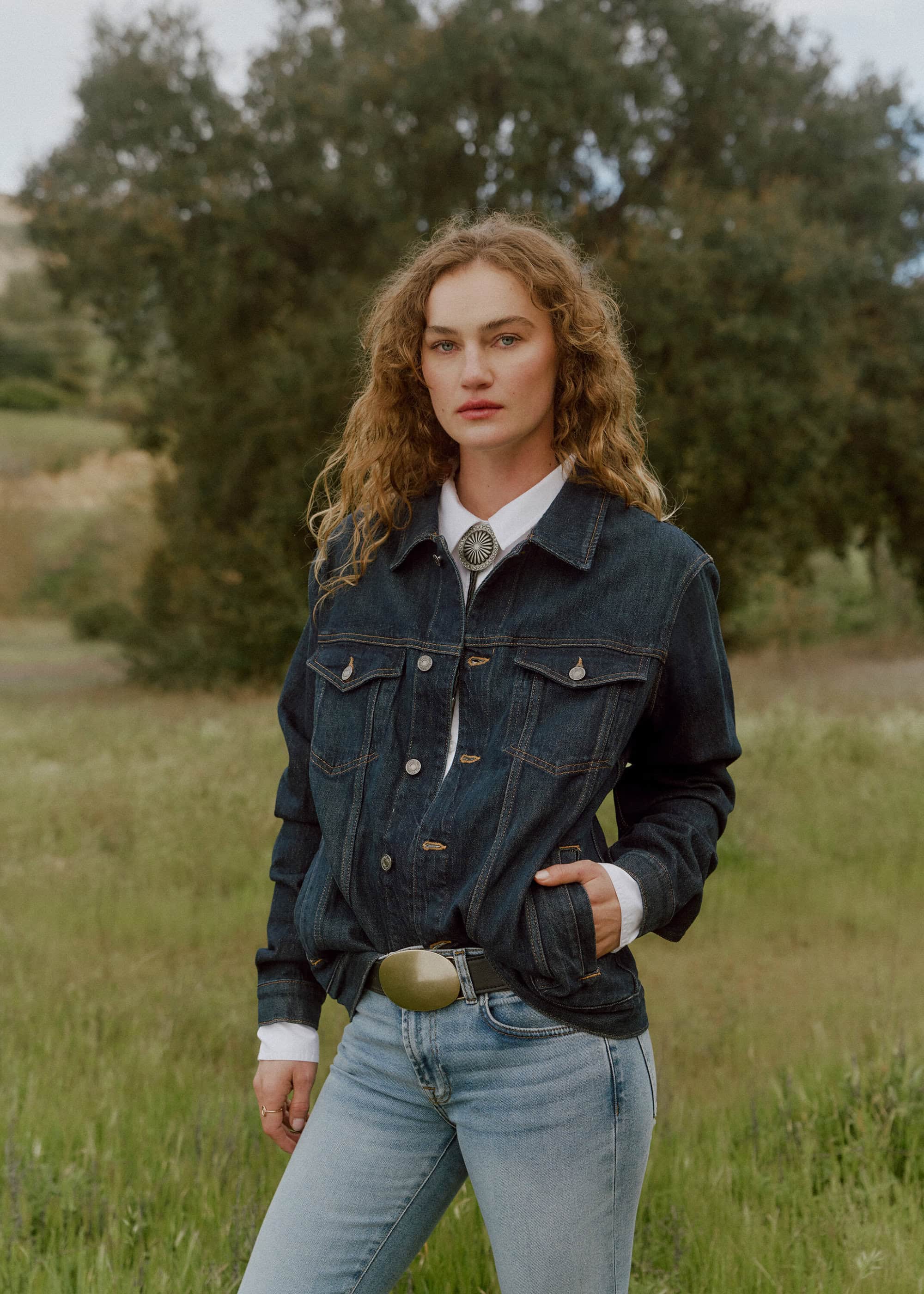 Woman with curly hair poses in denim jacket and jeans in a grassy field. Outdoor fashion shot with natural light and backdrop of trees.