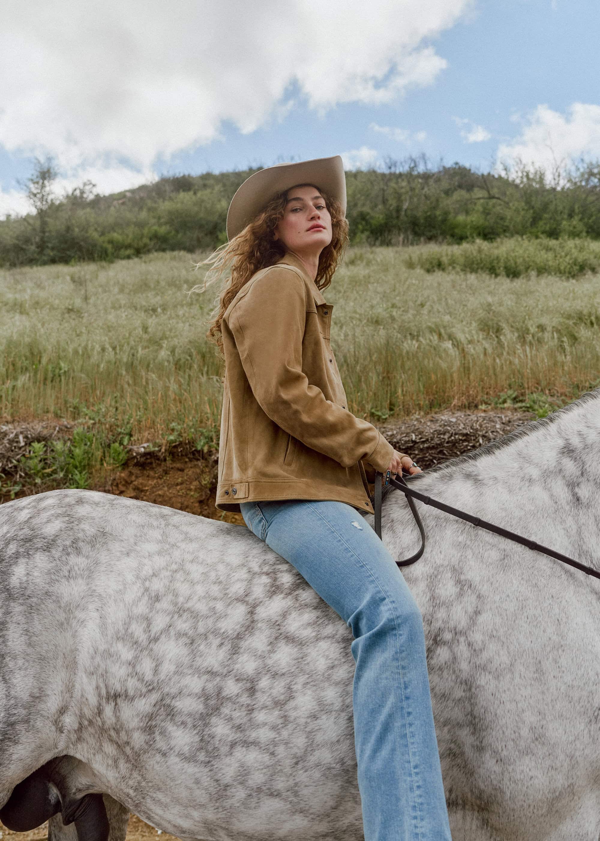 A woman with curly hair wearing a cowboy hat rides a dappled grey horse in a field under a cloudy sky, in a medium shot.