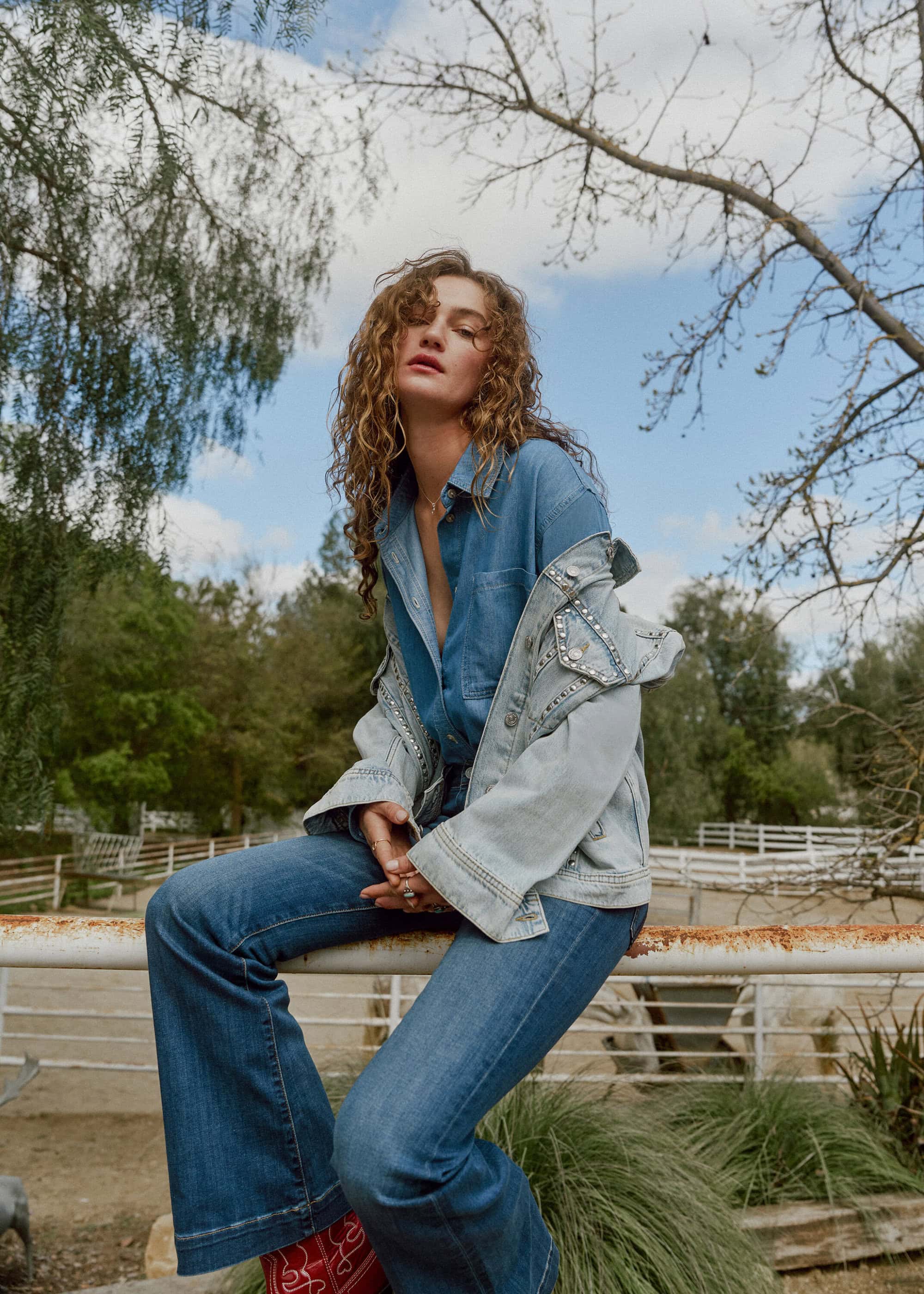 A woman in a denim outfit sits on a metal railing in an outdoor setting with trees and sky in the background, showcasing a casual style.