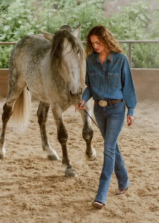 A woman walks with a grey horse on a leash in an indoor riding arena with a sandy surface and wooden fencing. They are the main focus.