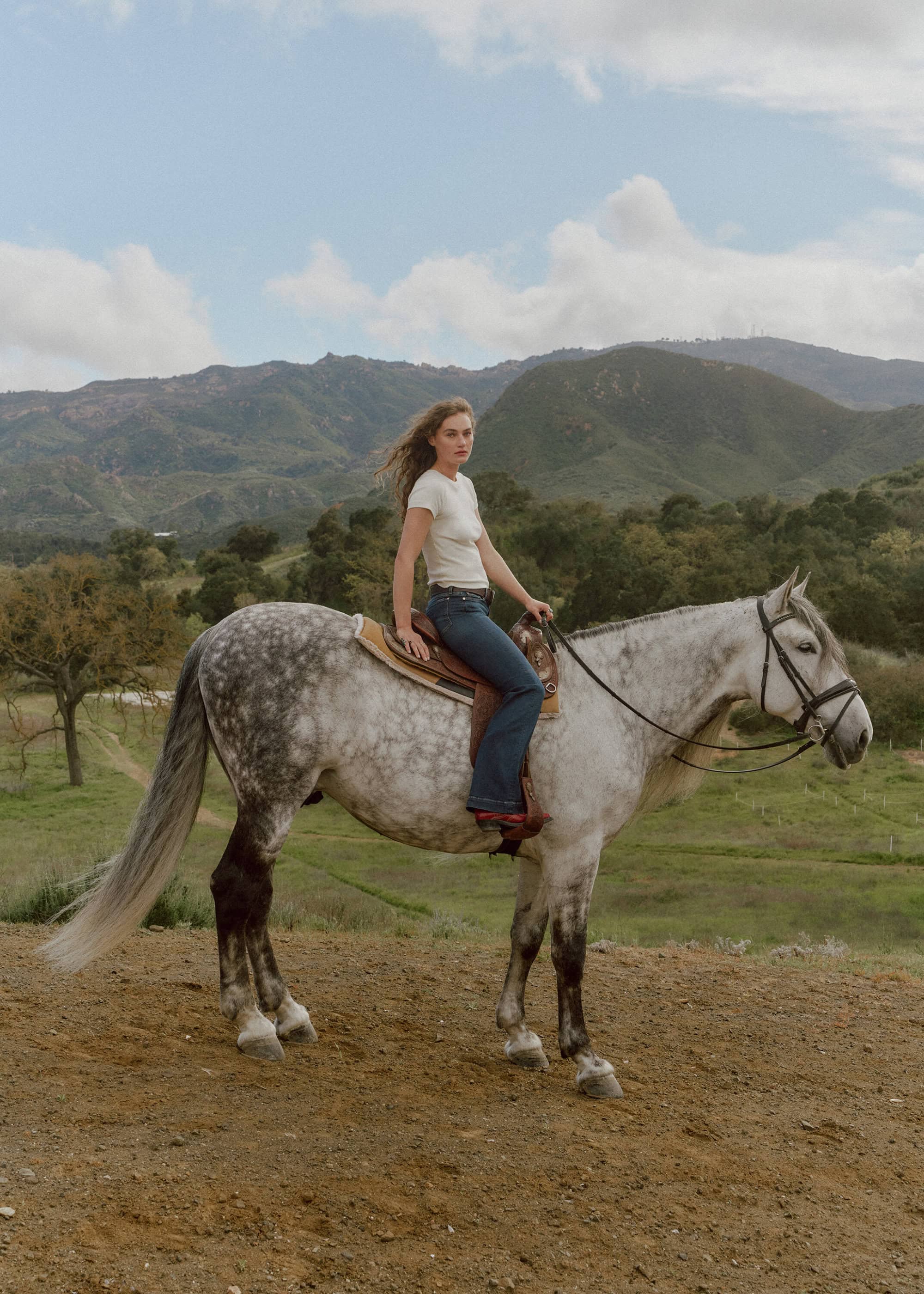 A woman rides a dappled gray horse in a grassy, rural setting with mountains in the background on a partly cloudy day.