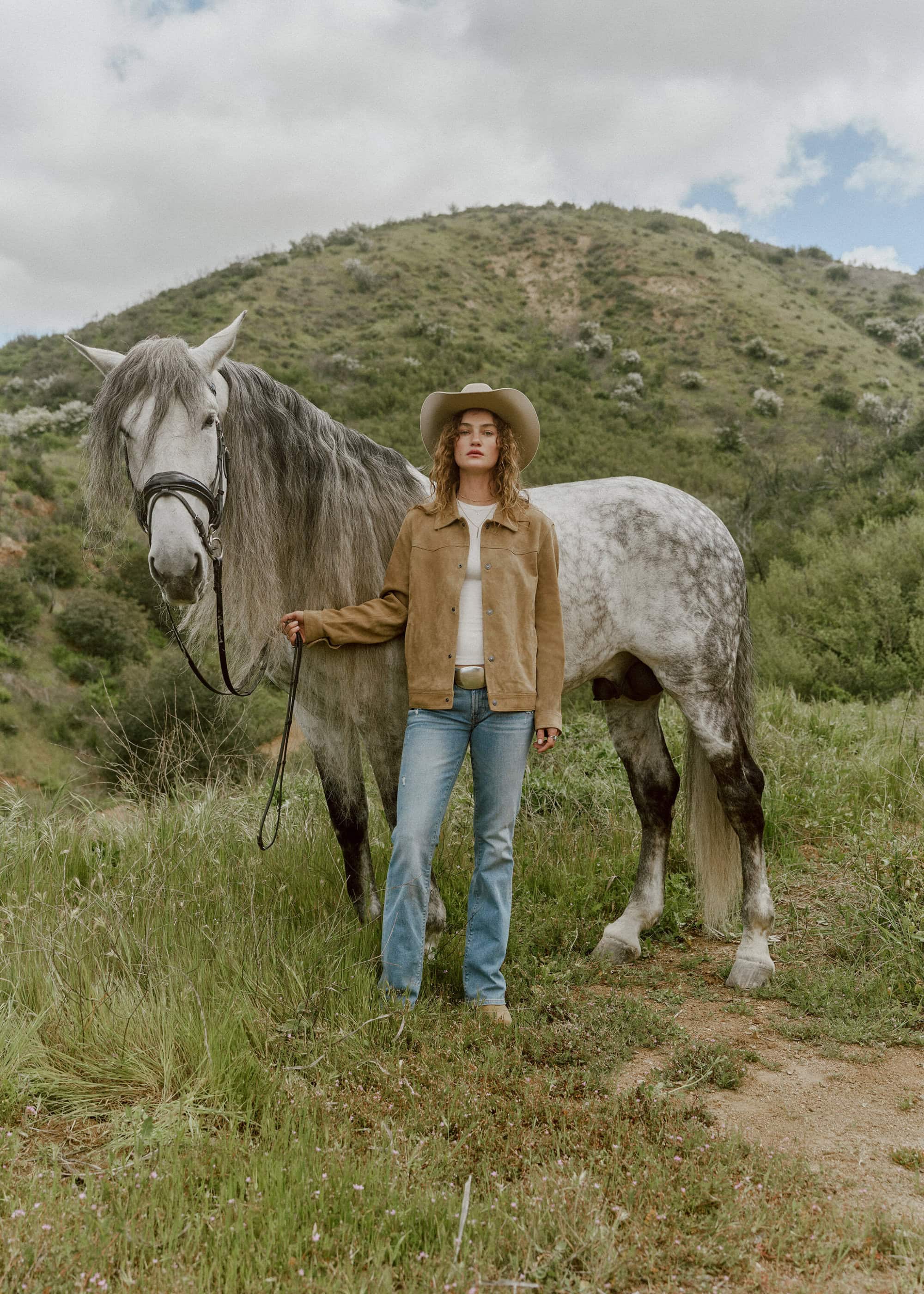 Woman in cowboy hat and jacket stands with a grey horse in a grassy field with a grassy hill and cloudy sky in the background.