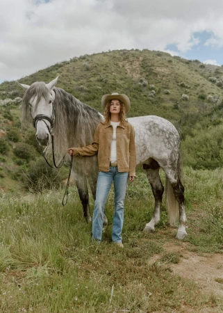 Woman in cowboy hat and jacket stands with a grey horse in a grassy field with a grassy hill and cloudy sky in the background.
