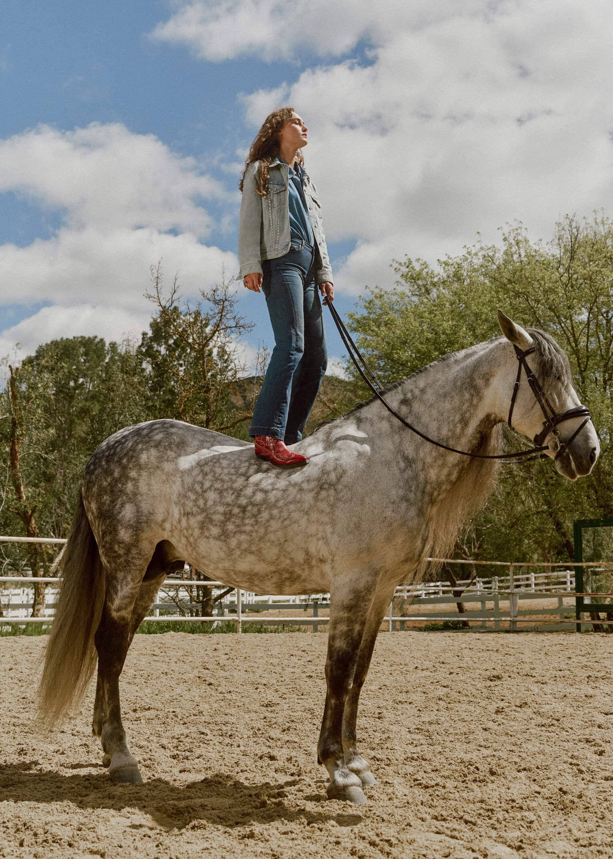 Woman stands on a dapple-gray horse in an outdoor paddock. She wears a denim outfit and red cowboy boots. Clouds fill the sky.