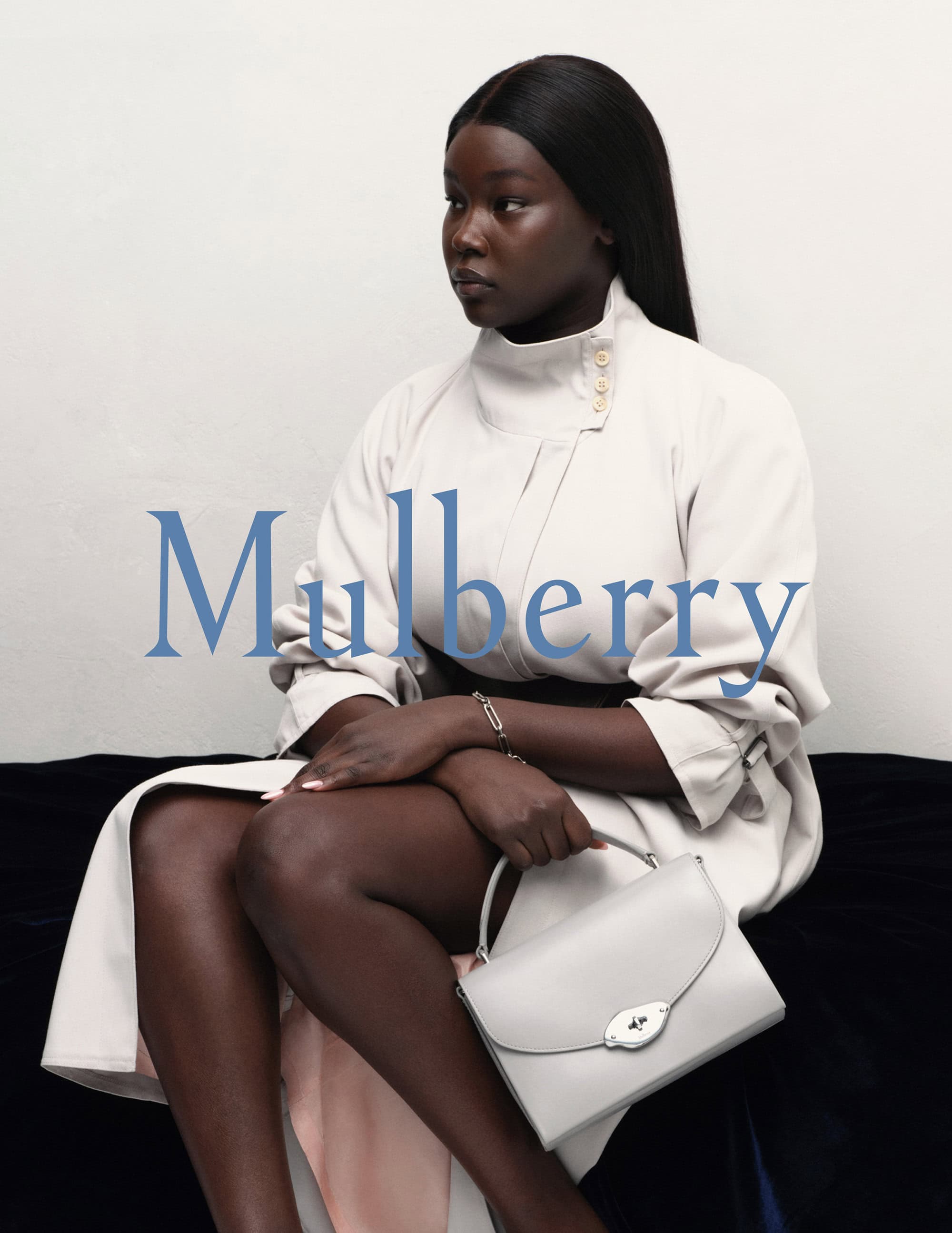 Elegant African-American woman posing with a light gray handbag in a fashion-forward cream-colored dress, seated against a neutral backdrop.
