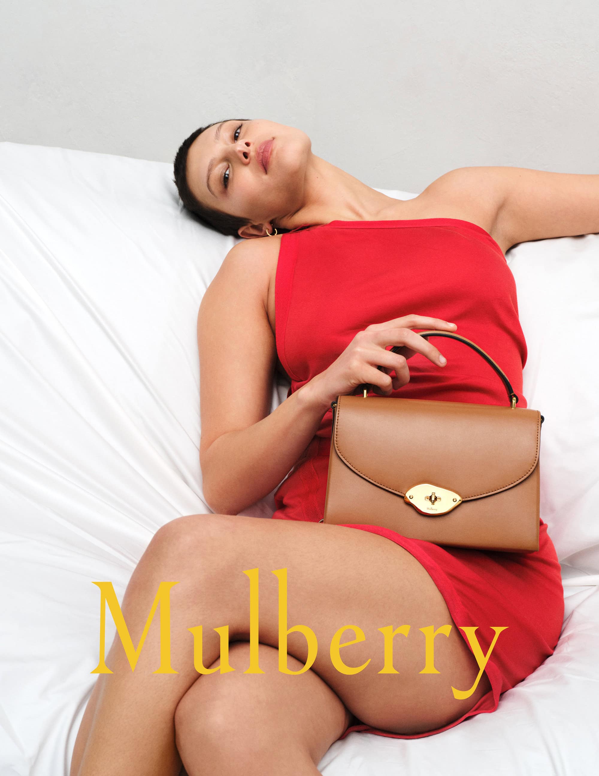 Woman poses in red dress, holding a tan leather satchel bag with gold hardware on white bedding, with a logo overlay.