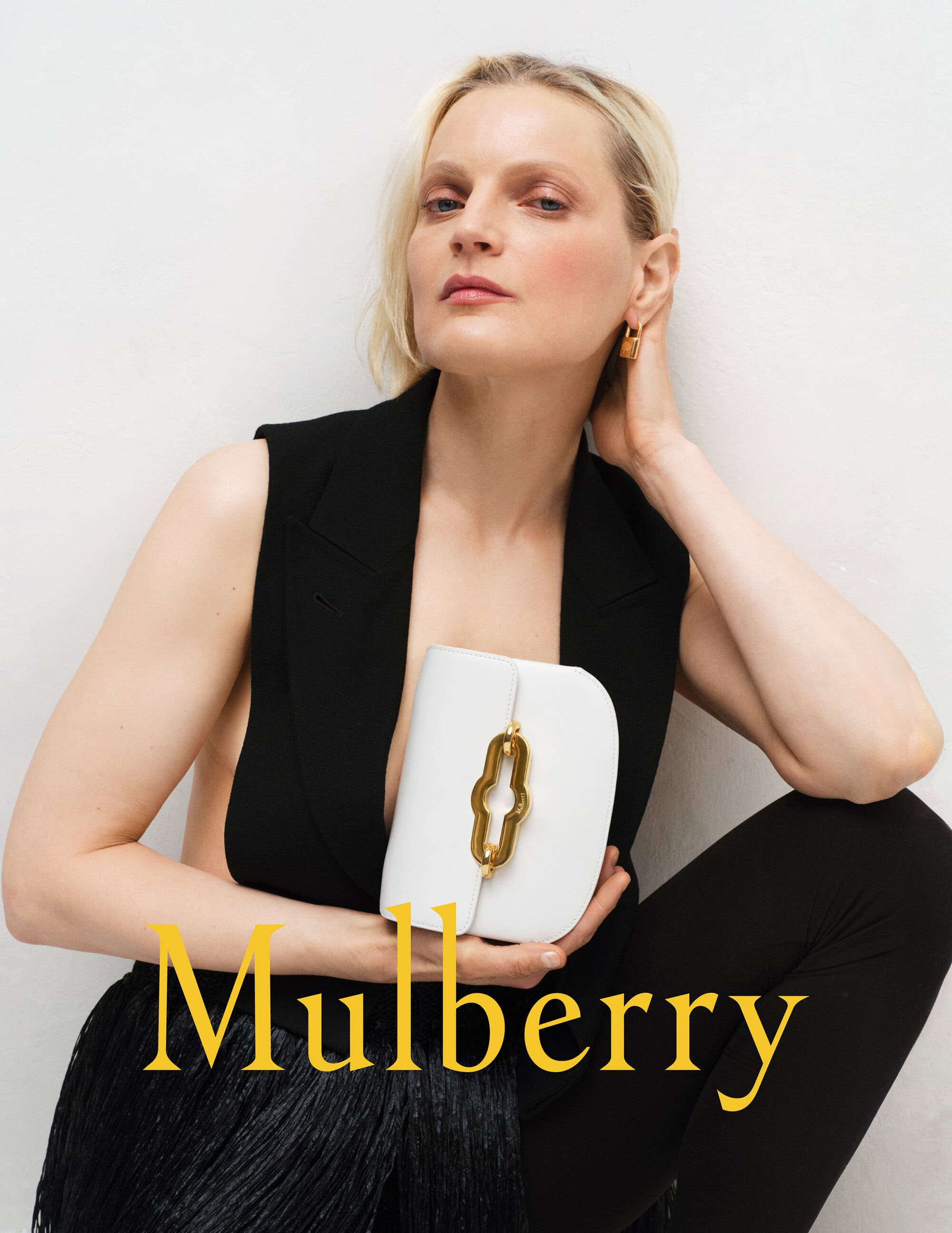 Fashion shot of a blonde model posing with a white leather Mulberry handbag, wearing a black vest top and black trousers against white backdrop.