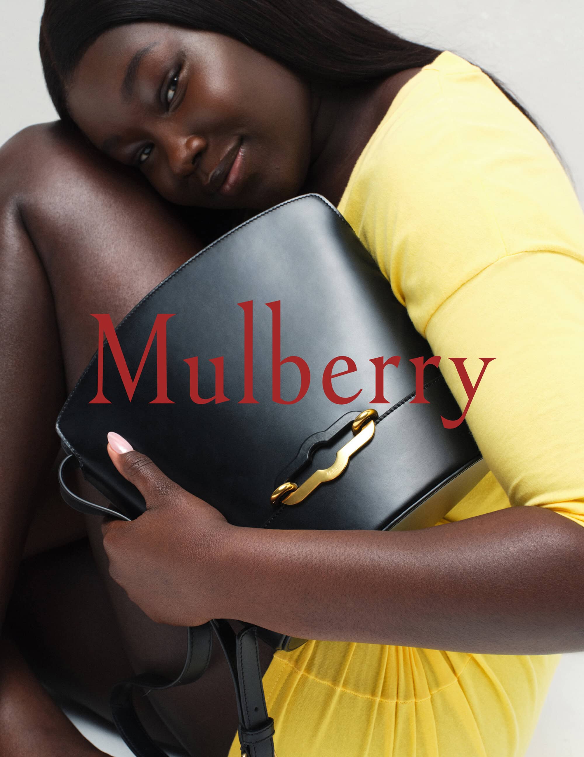 Elegant dark-skinned woman poses with a black leather Mulberry handbag, wearing a yellow dress, showcasing fashion and luxury.