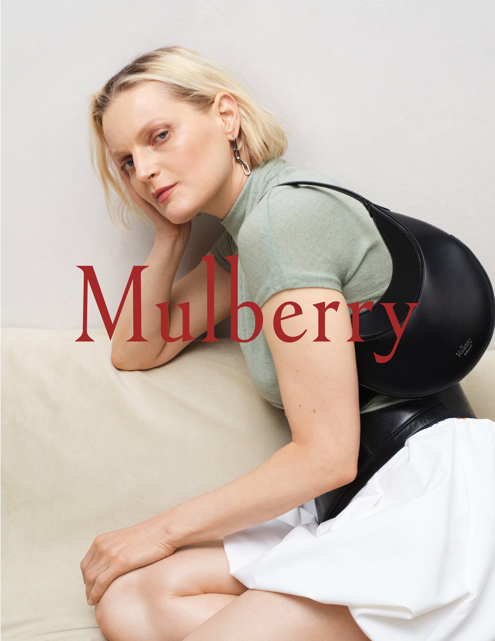 A blonde woman leaning against a wall wearing a sage green top and a white skirt while holding a black leather Mulberry shoulder bag.