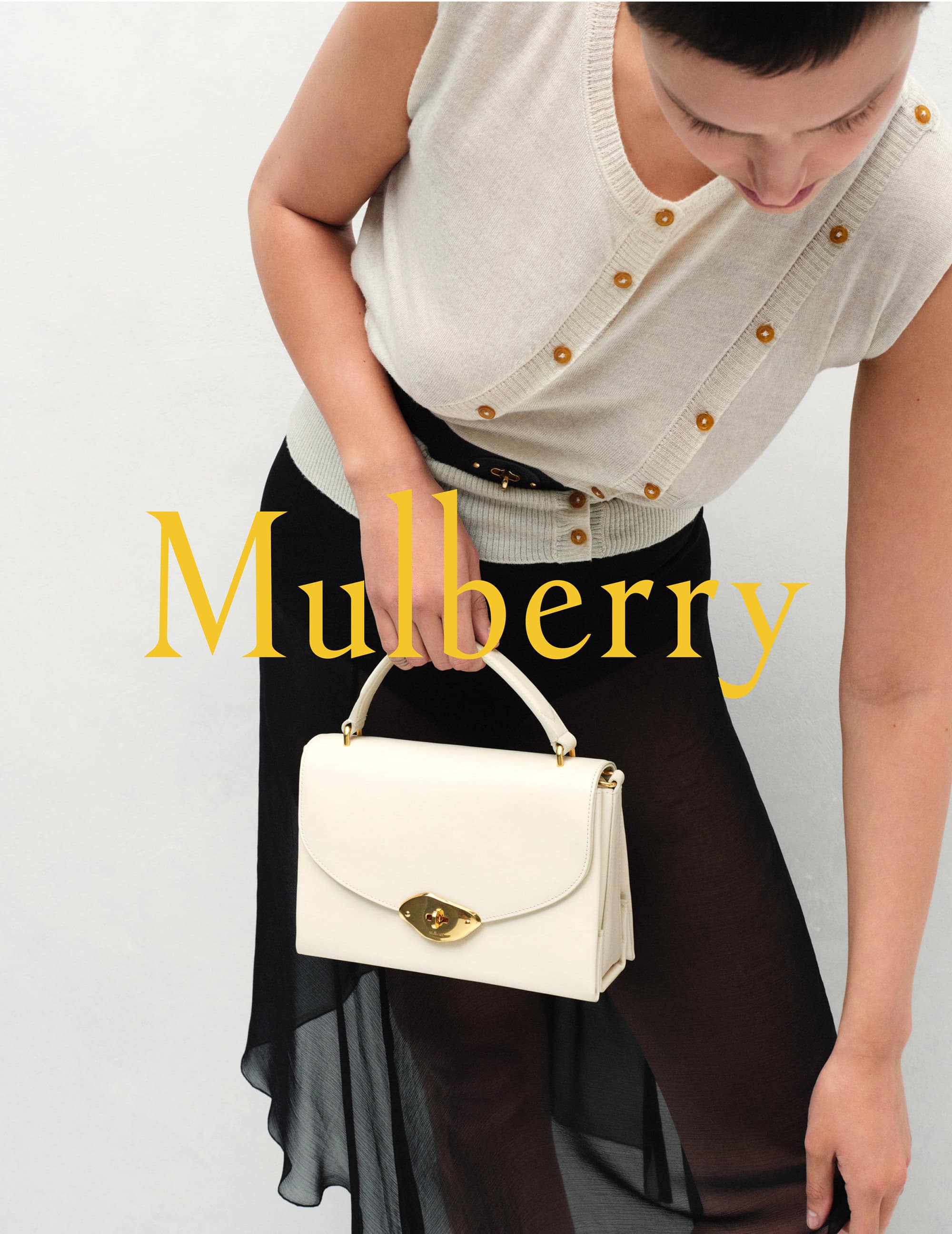 Fashionable woman showcases a cream-colored leather handbag with a gold clasp, paired with a light button-down sweater and sheer black skirt.