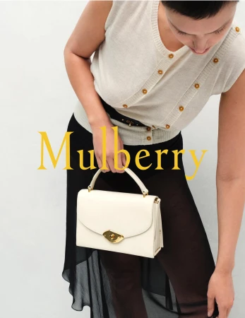 Fashionable woman showcases a cream-colored leather handbag with a gold clasp, paired with a light button-down sweater and sheer black skirt.