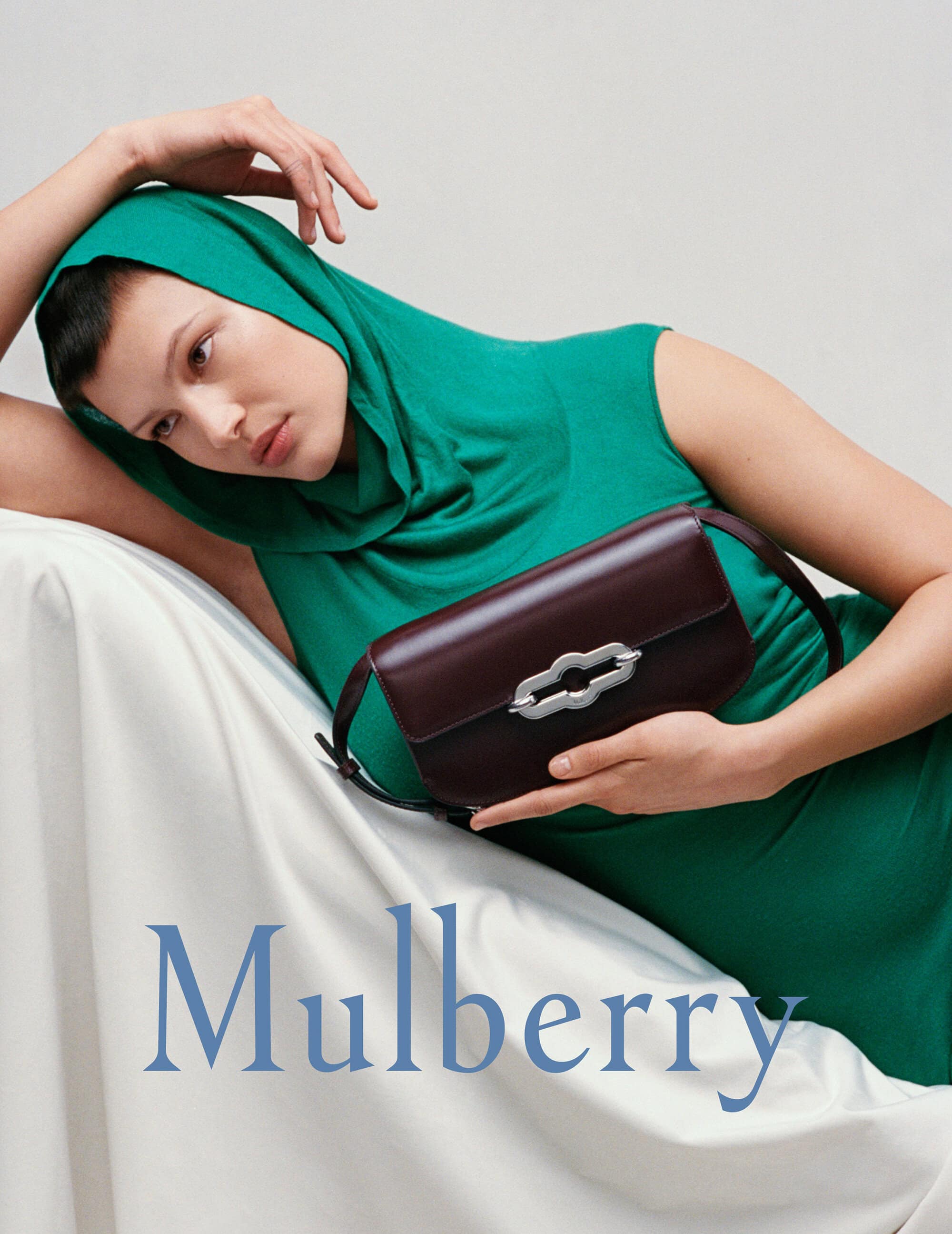 Brunette adult woman in a green dress reclines while holding a dark brown leather handbag with a silver clasp in a studio setting.