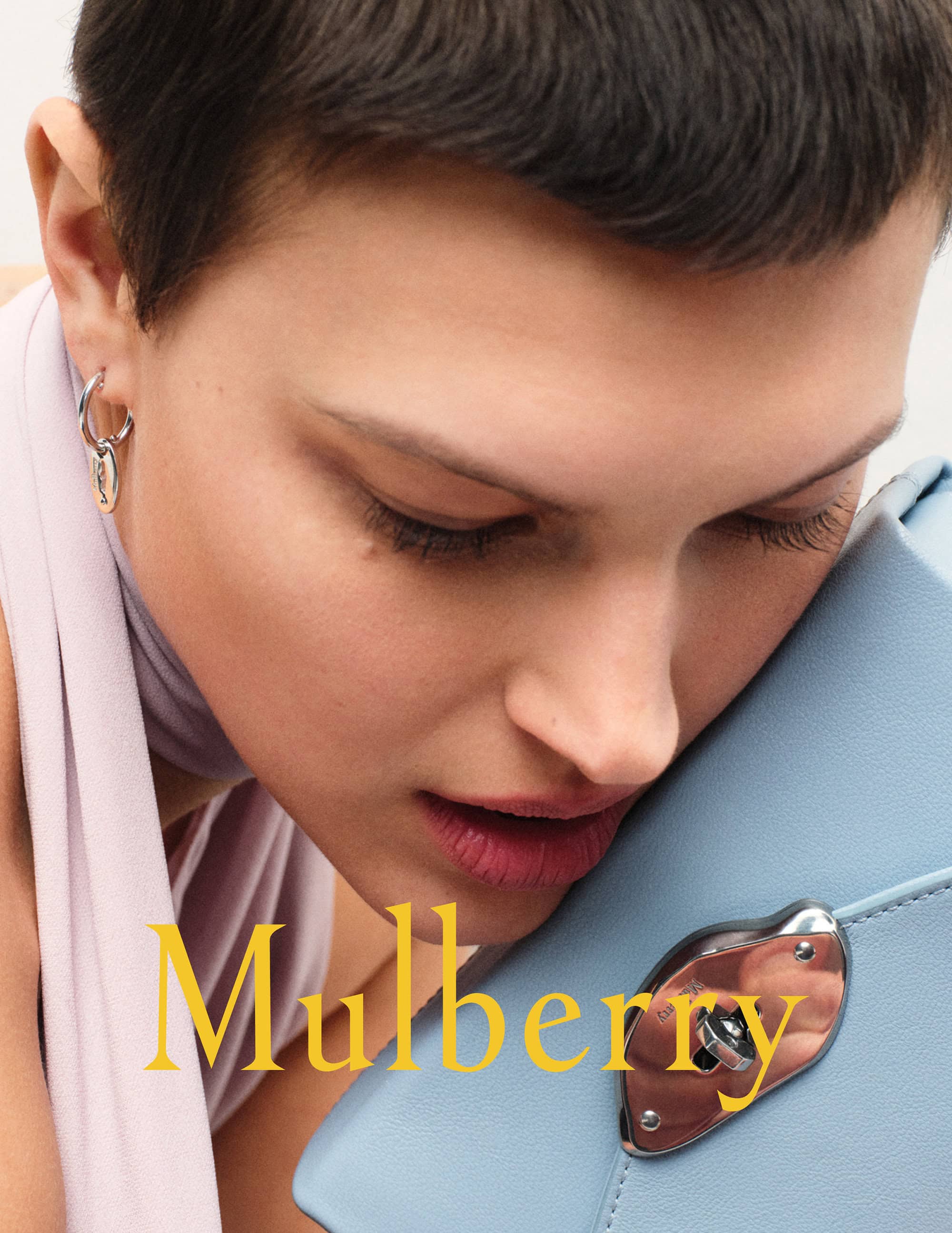 Close-up of a woman with short dark hair, wearing hoop earrings and a lavender scarf, resting her cheek on a light blue purse.