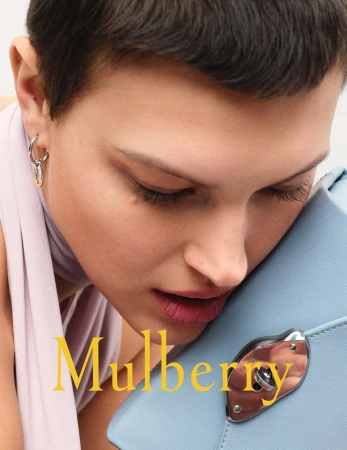 Close-up of a woman with short dark hair, wearing hoop earrings and a lavender scarf, resting her cheek on a light blue purse.