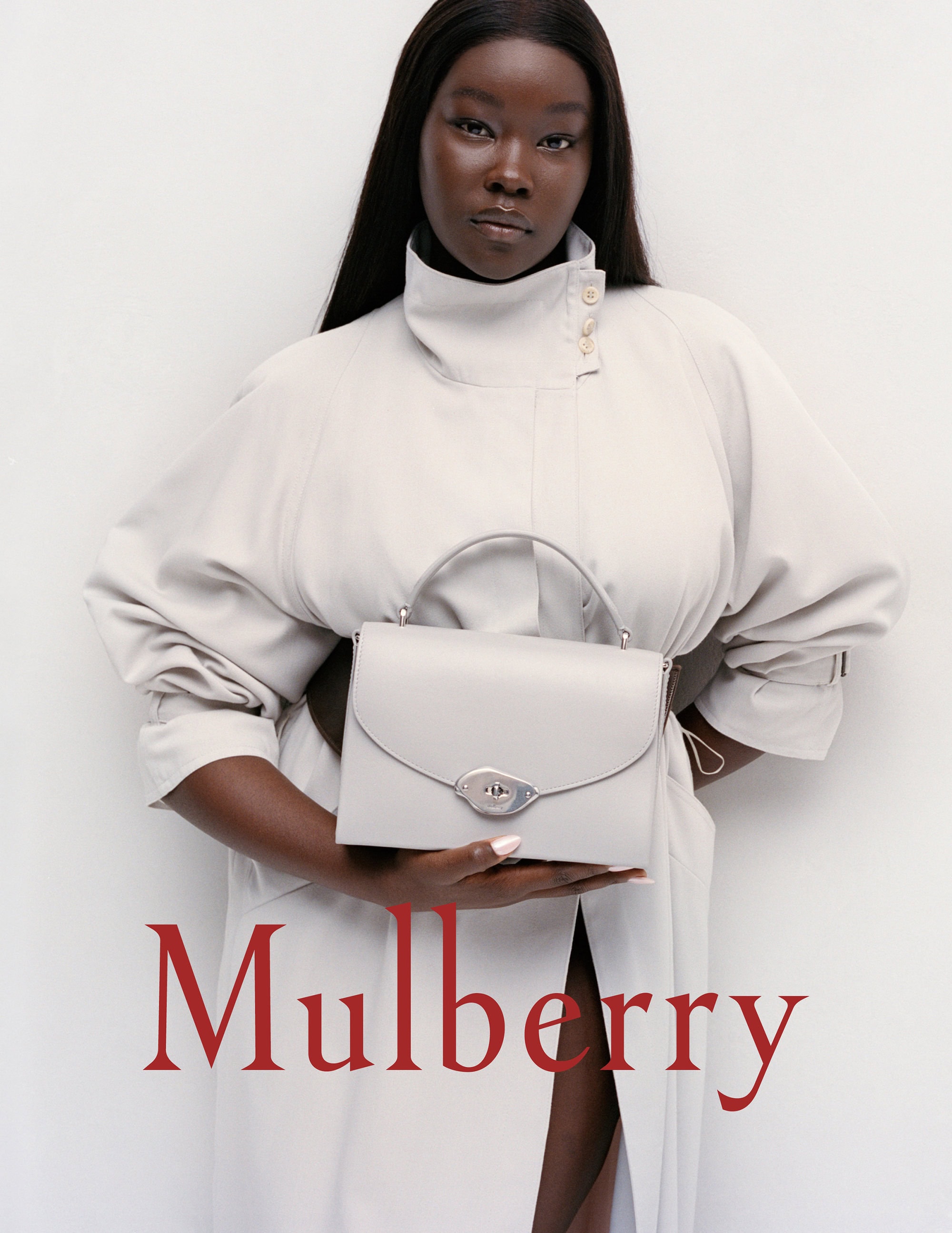 Elegant studio shot of a Black female model holding a light grey Mulberry handbag, wearing a light grey trench coat against white backdrop.
