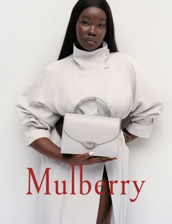 Elegant studio shot of a Black female model holding a light grey Mulberry handbag, wearing a light grey trench coat against white backdrop.