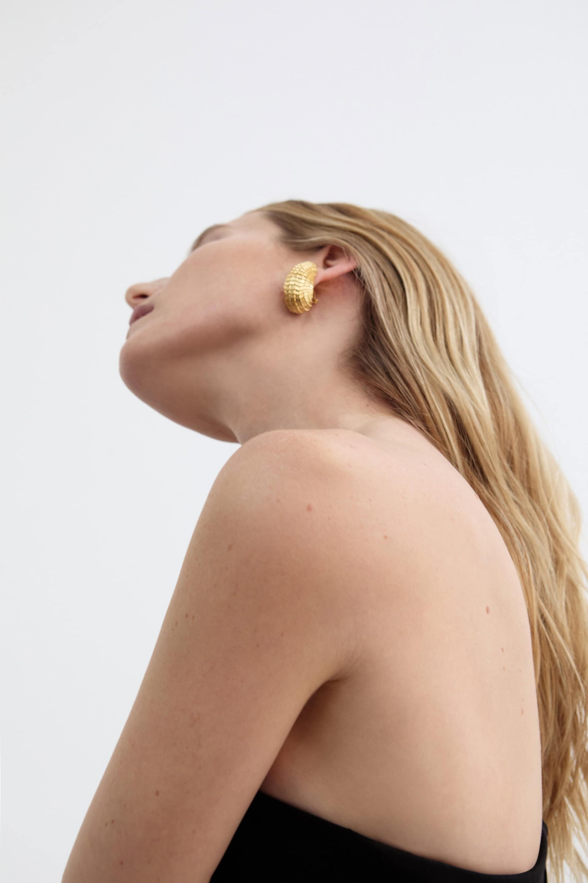 Model Dree Hemingway tilting her head back wearing a textured gold earring and black top against a white seamless background.