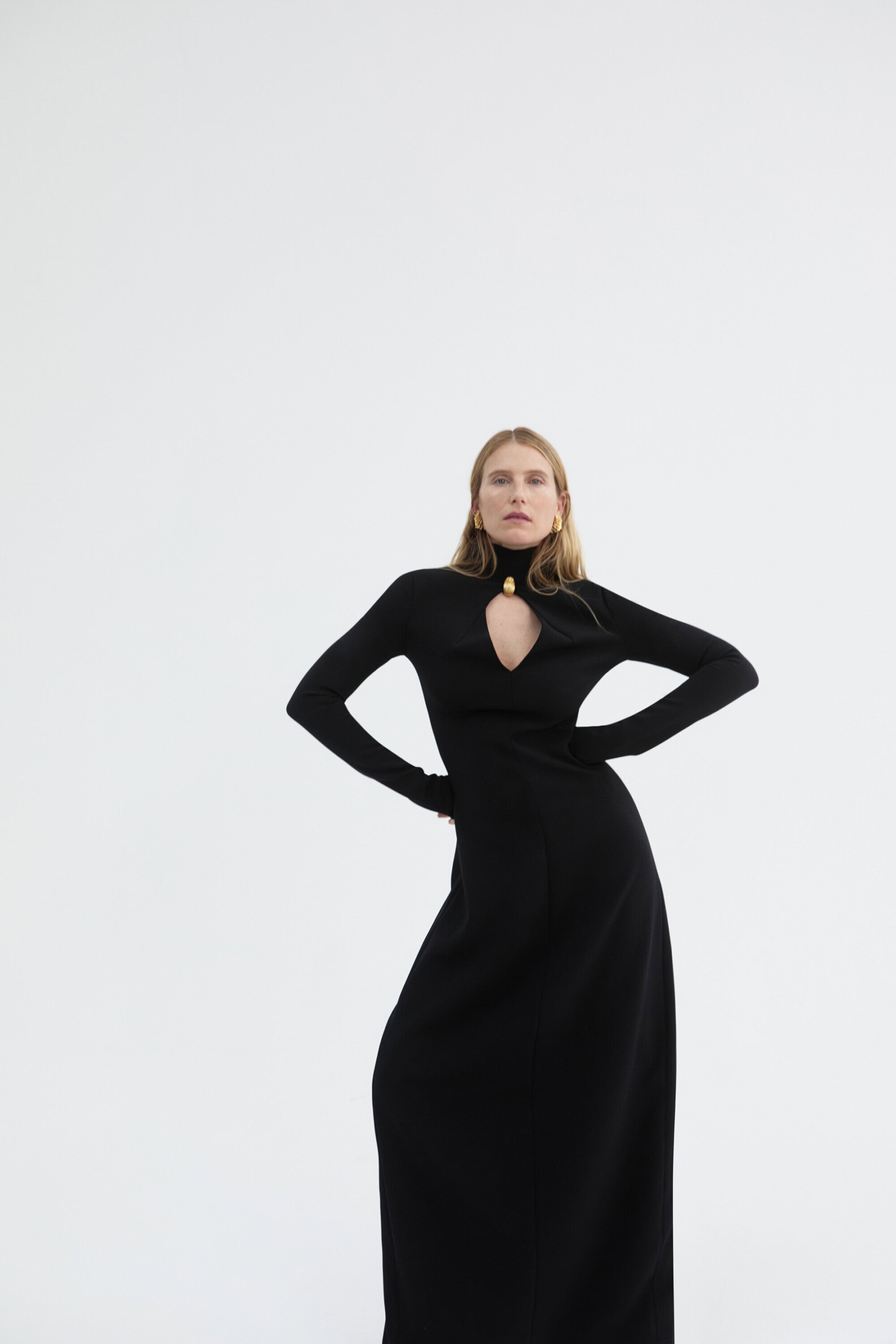 Elegant woman in a long-sleeved black maxi dress with a gold pendant posing in a white studio setting with hands on her hips.
