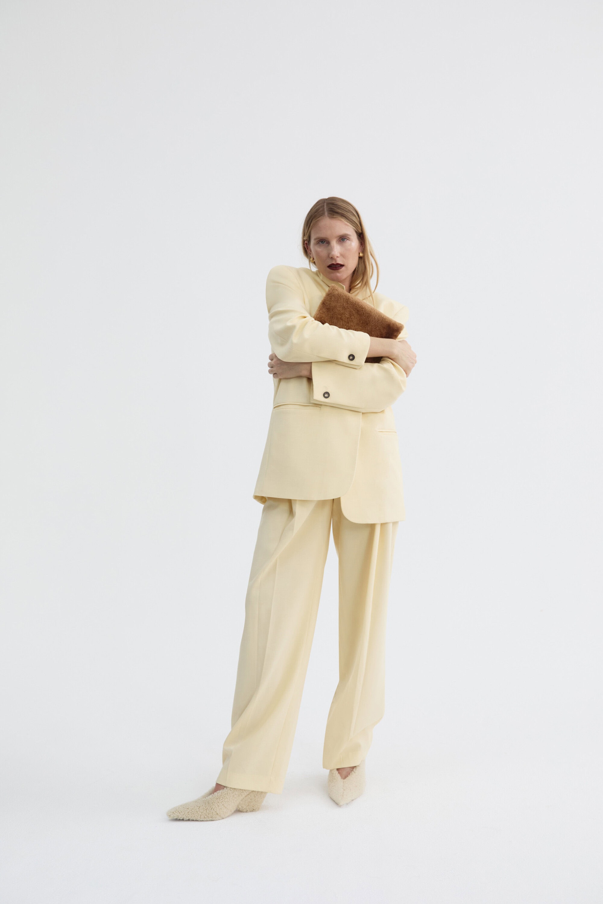 A woman in a cream-colored suit hugs a brown clutch in a studio setting against a white backdrop in a full shot view.