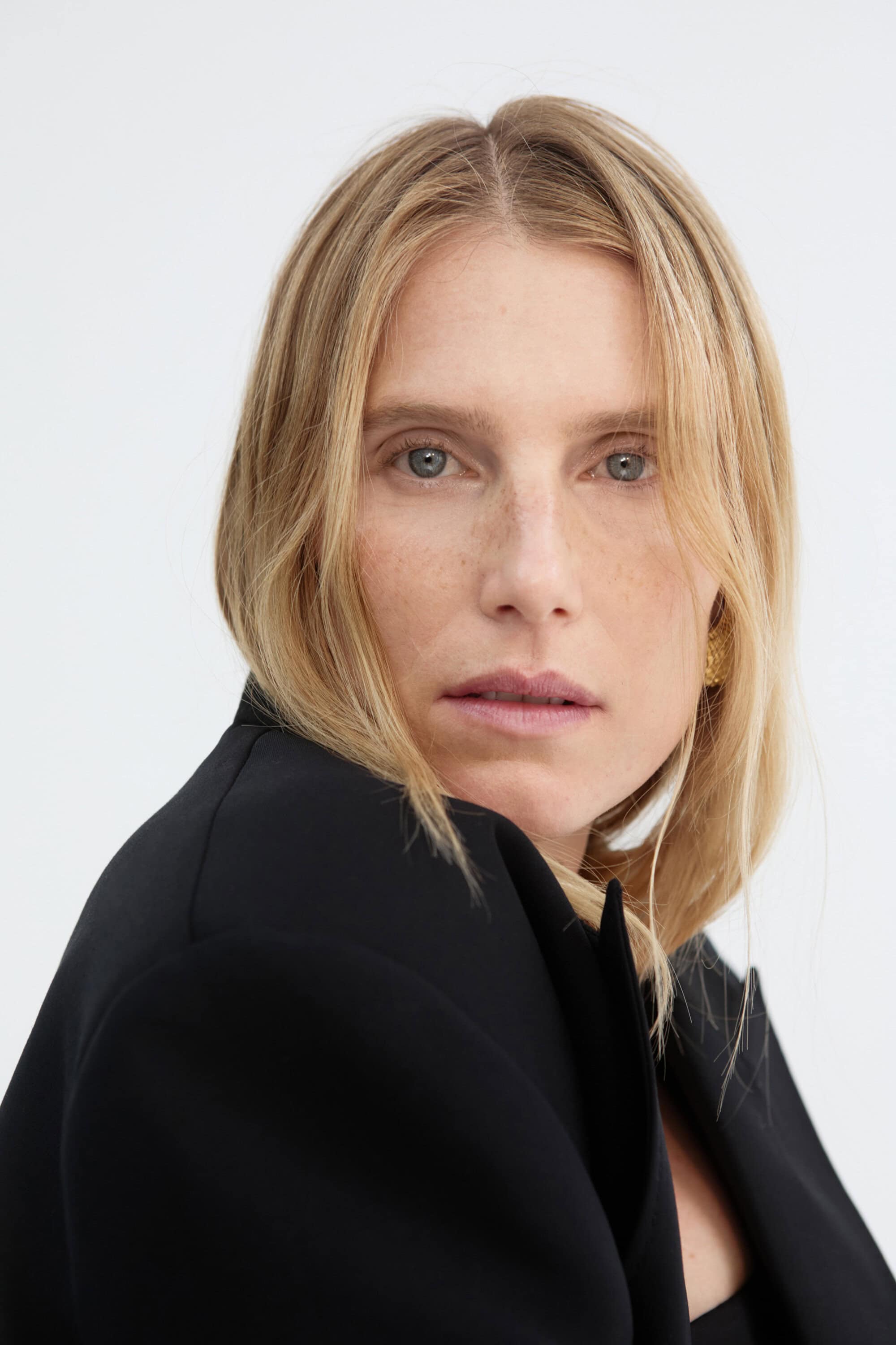 Blonde woman in a black blazer poses against a white background in a close-up shot for a fashion or beauty portrait.