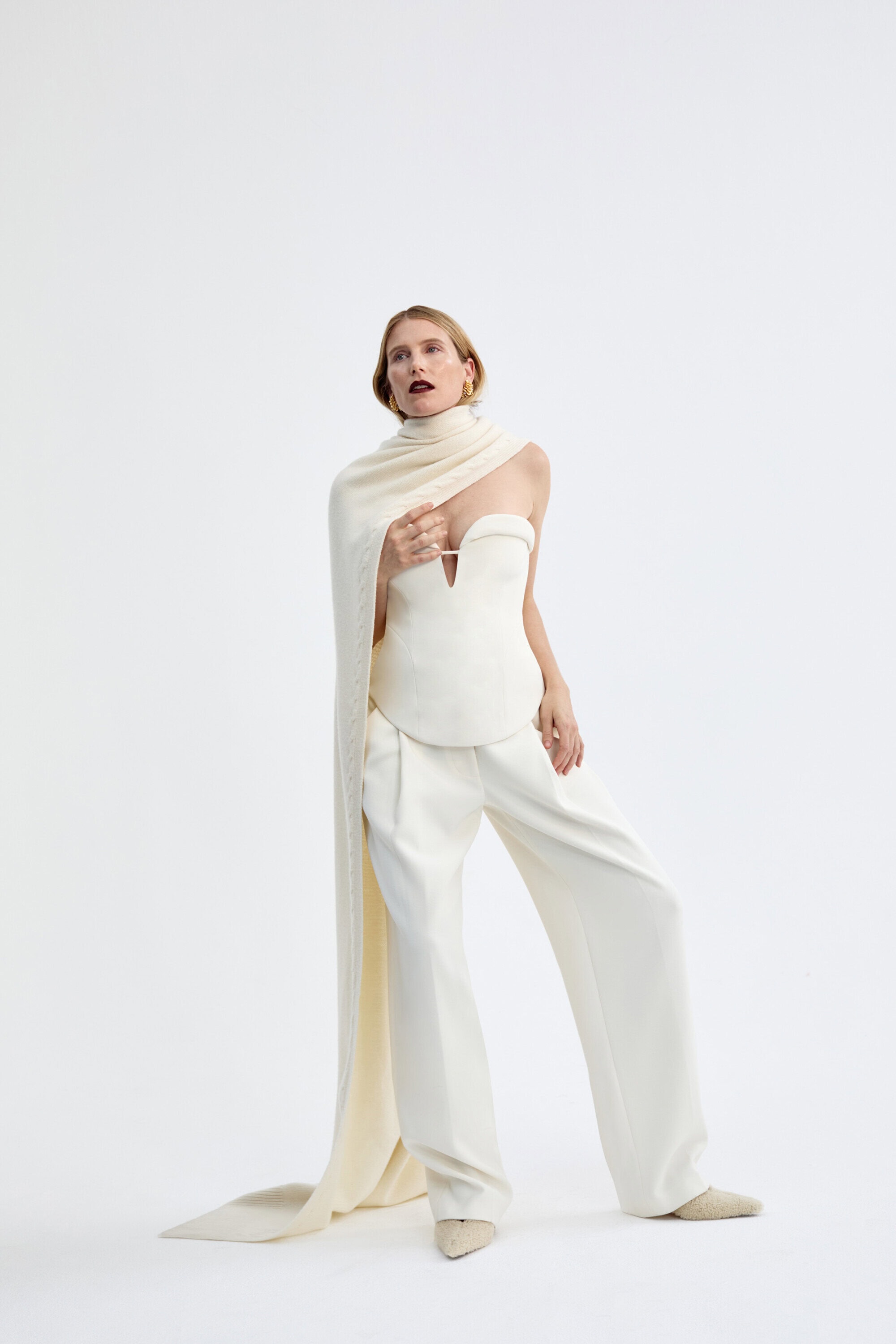 A fashion model wearing an off-white strapless corset top, wide-leg trousers, and a long draped scarf against a plain white studio backdrop.