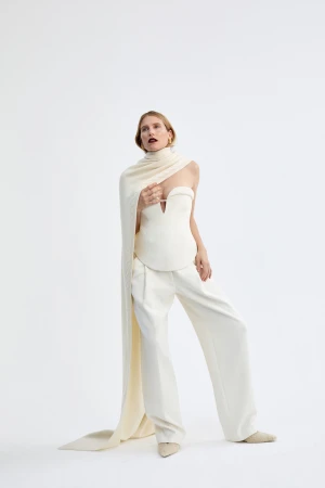 A fashion model wearing an off-white strapless corset top, wide-leg trousers, and a long draped scarf against a plain white studio backdrop.
