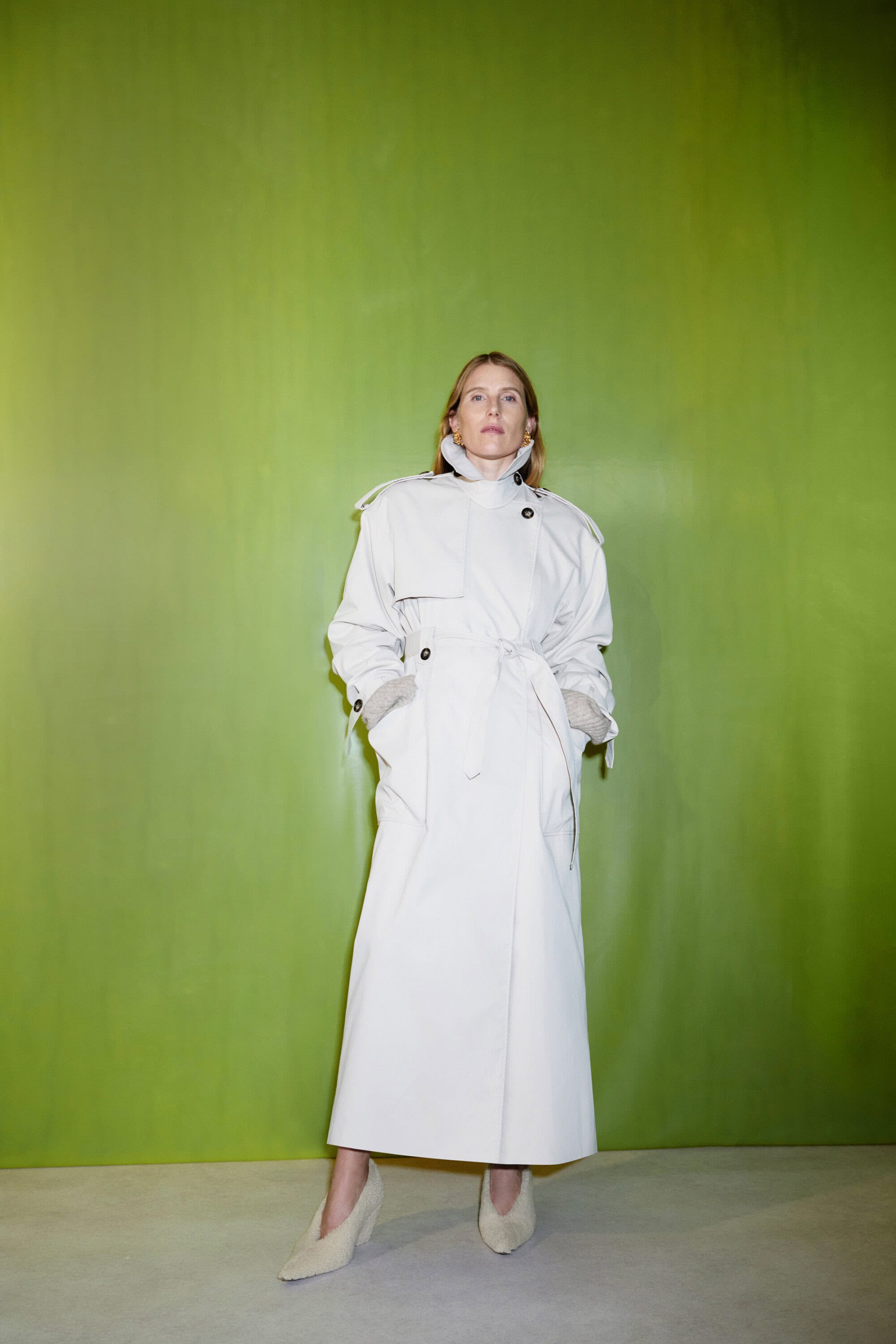 Woman in a trench coat stands against a green wall. She wears neutral shoes and her hands are in her pockets. Studio setting.
