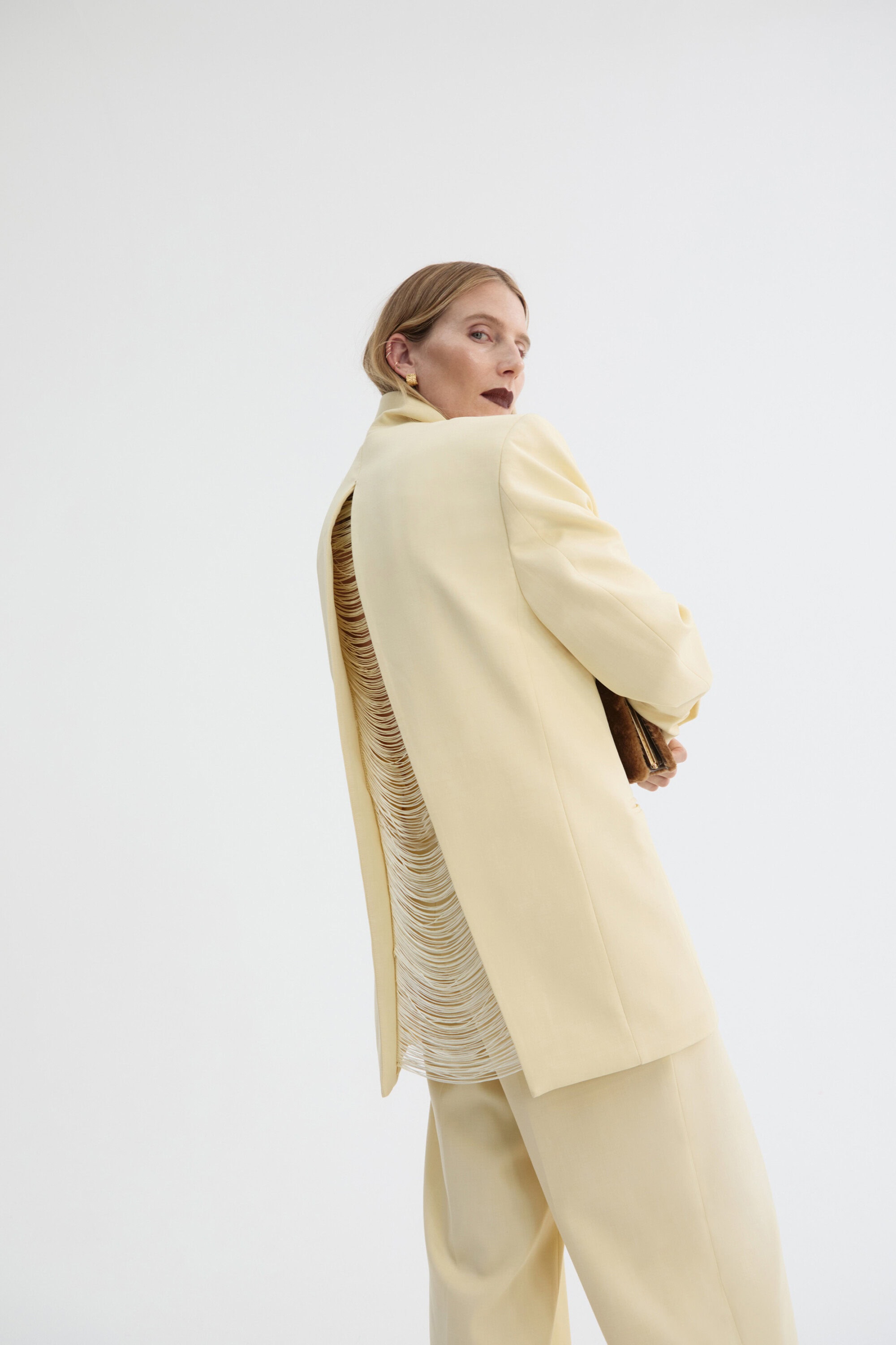 Woman in a light yellow suit with fringe detail, standing against a white backdrop, looking over her shoulder.