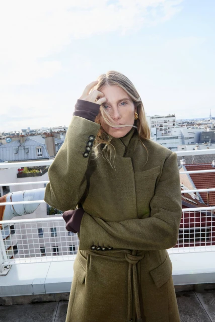 Adult woman in a green trench coat stands on a rooftop with a city skyline in the background on a sunny day, fashion forward.