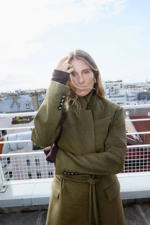 Adult woman in a green trench coat stands on a rooftop with a city skyline in the background on a sunny day, fashion forward.