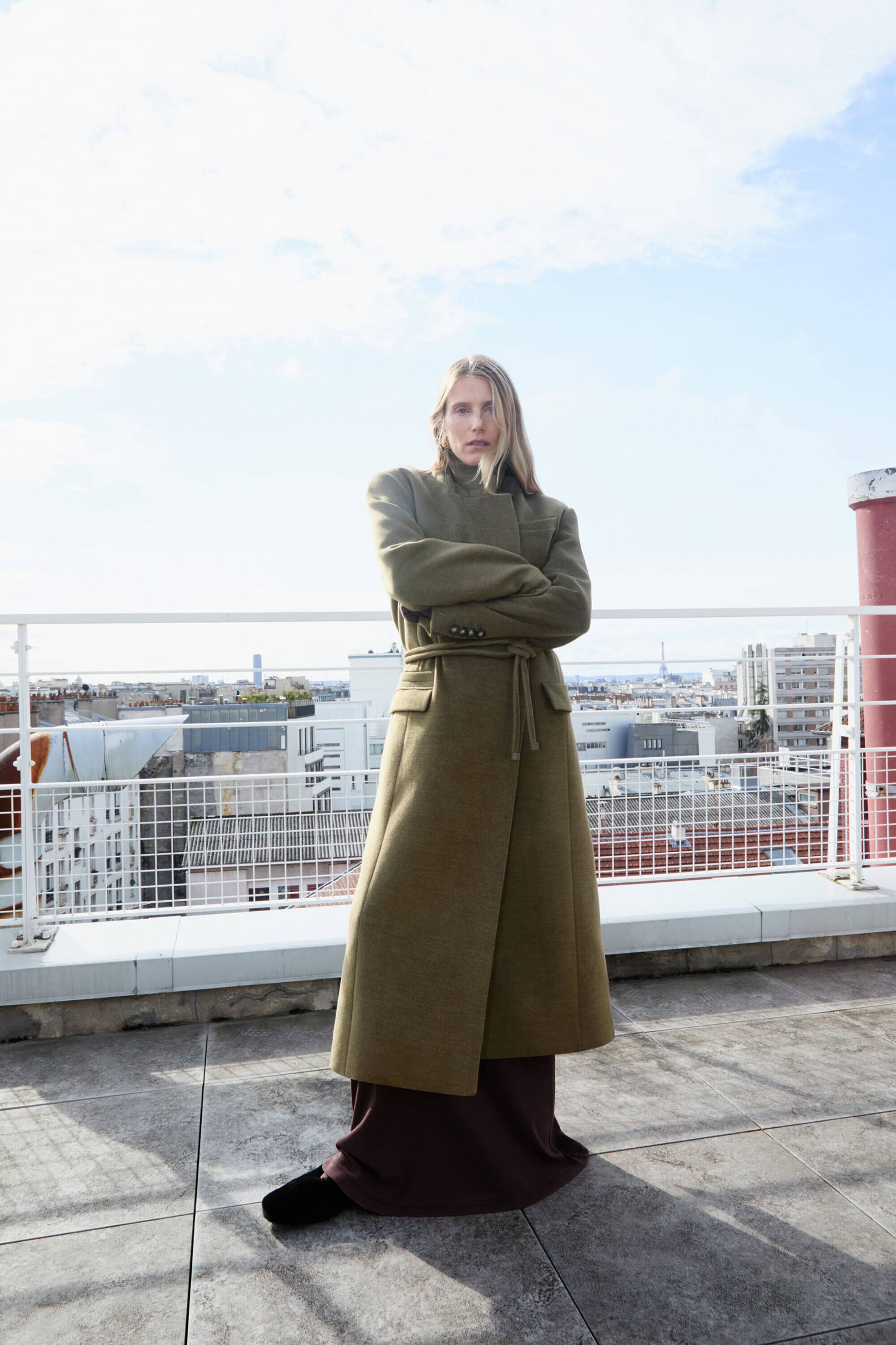 Model wearing a long olive green belted coat standing on a concrete rooftop with city views