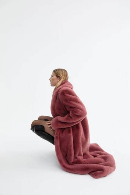 Fashion shot of a woman in a long pink faux-fur coat and high black boots, crouching indoors against a white backdrop.