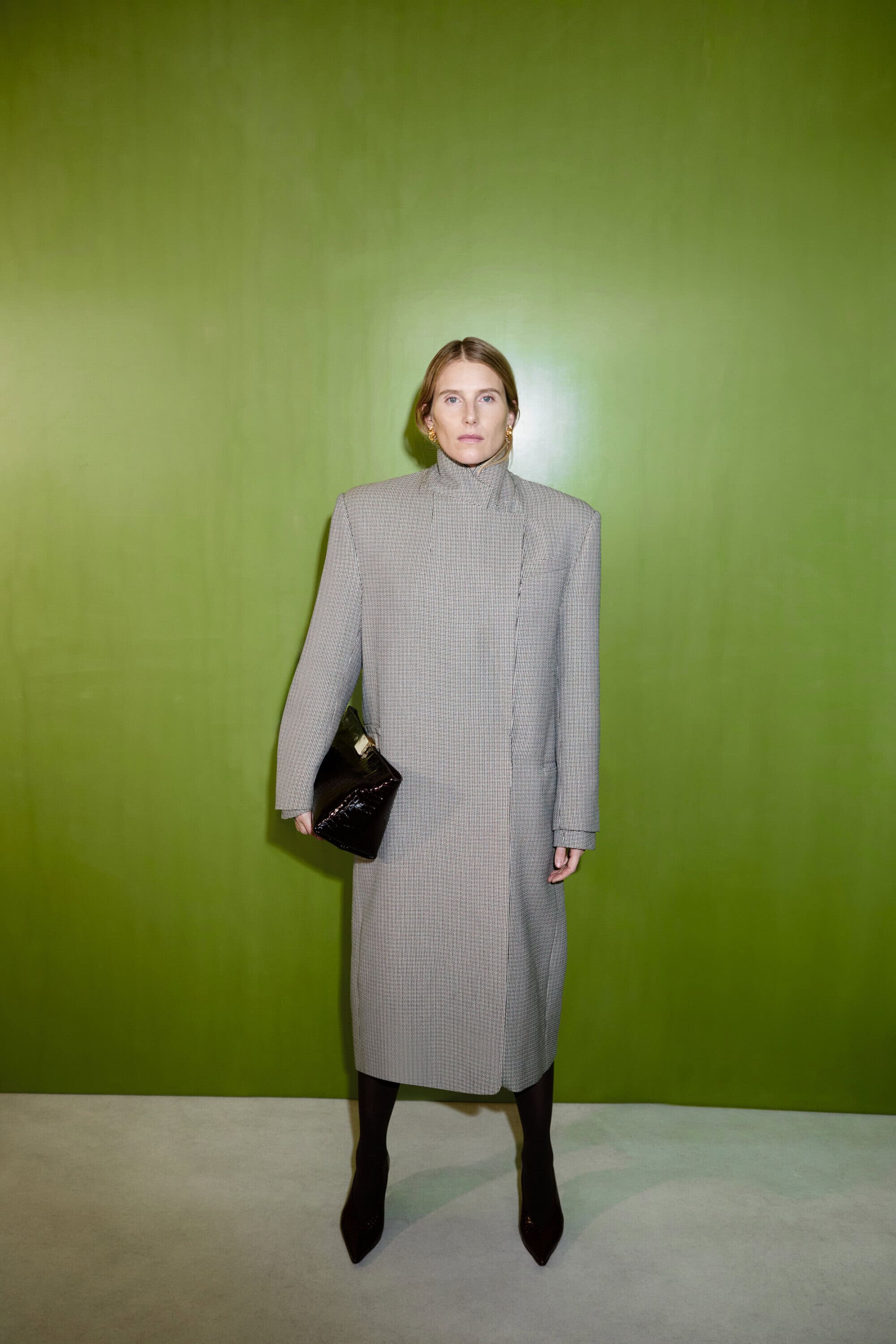 Woman in a houndstooth coat poses in front of a lime green wall, holding a black clutch and wearing pointy-toe boots for a fashion portrait.