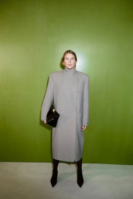 Woman in a houndstooth coat poses in front of a lime green wall, holding a black clutch and wearing pointy-toe boots for a fashion portrait.