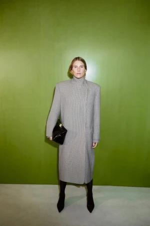 Woman in a houndstooth coat poses in front of a lime green wall, holding a black clutch and wearing pointy-toe boots for a fashion portrait.