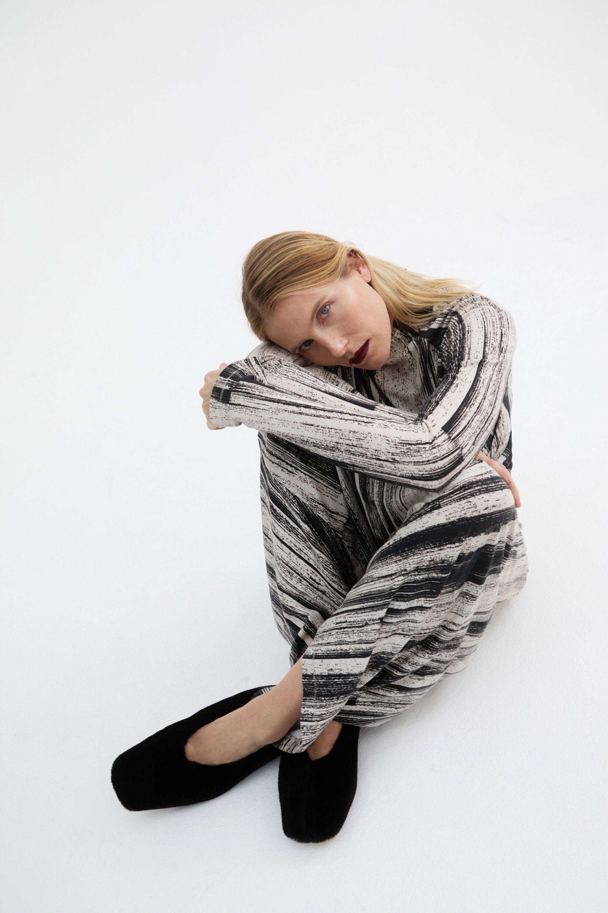 Blonde woman in a patterned long-sleeve top and pants sitting on a white seamless background in a relaxed pose wearing black slippers.
