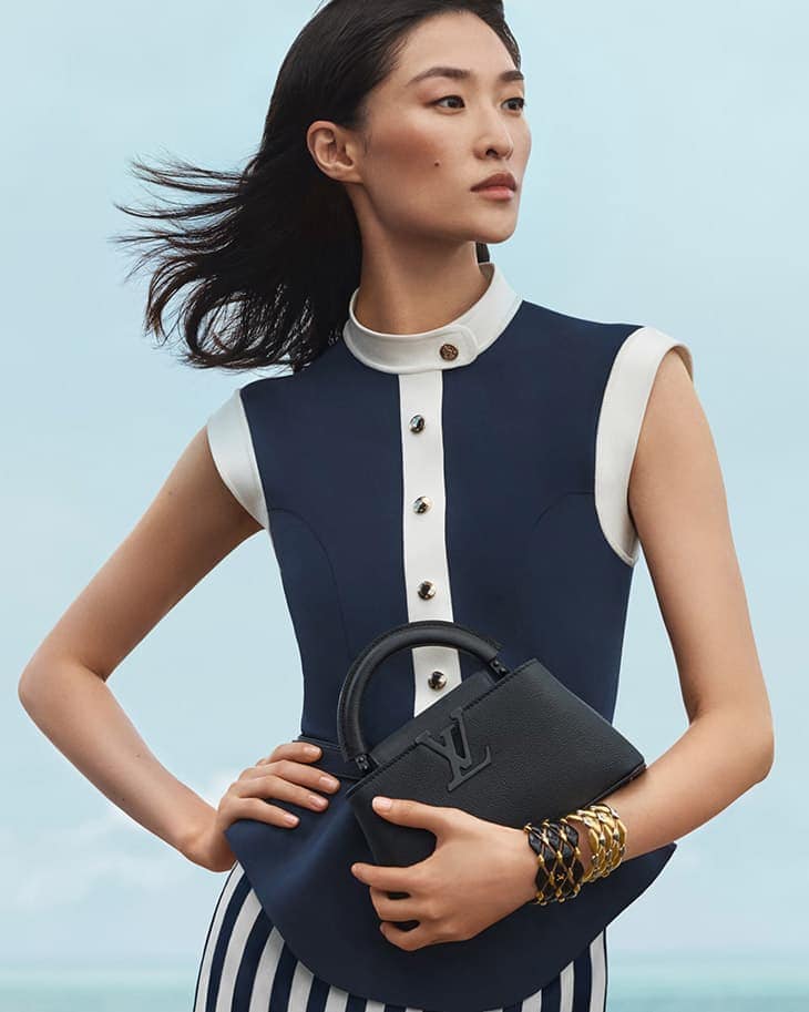 Elegant Asian woman in navy and white dress holding black Louis Vuitton handbag with bracelets, against a light blue background.