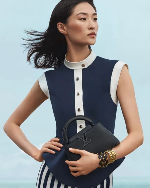 Elegant Asian woman in navy and white dress holding black Louis Vuitton handbag with bracelets, against a light blue background.