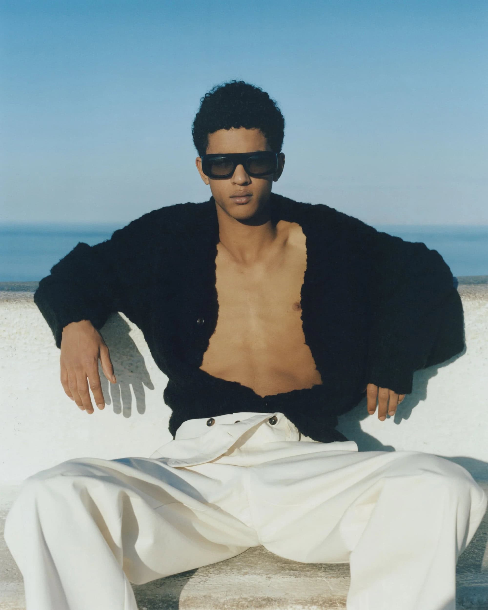 Man in sunglasses and black cardigan poses outdoors in sunlight with a horizon background of sky and sea wearing off-white pants.