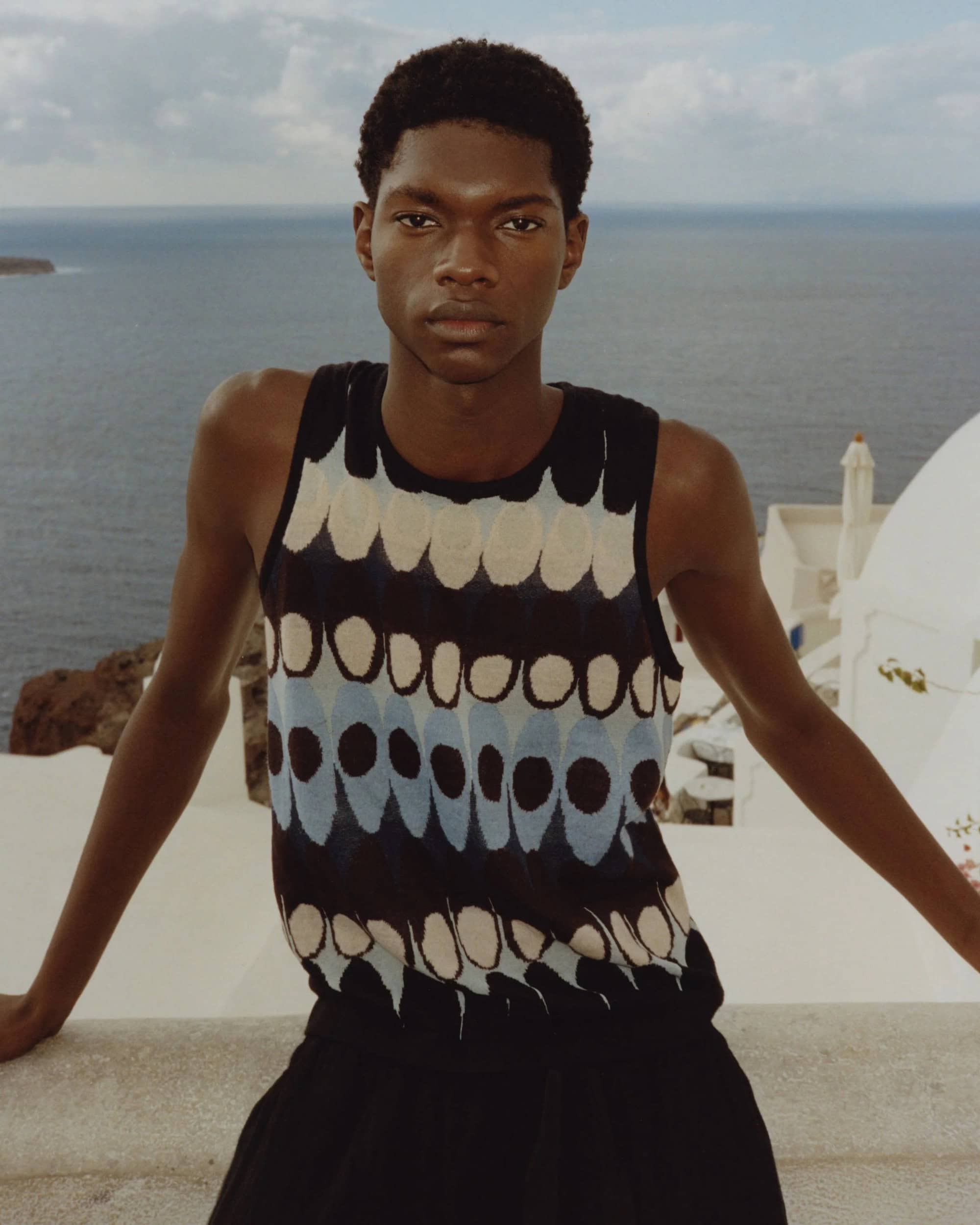 Fashion shot of a Black male model in a patterned top, posing outdoors with the ocean and white buildings in the background.