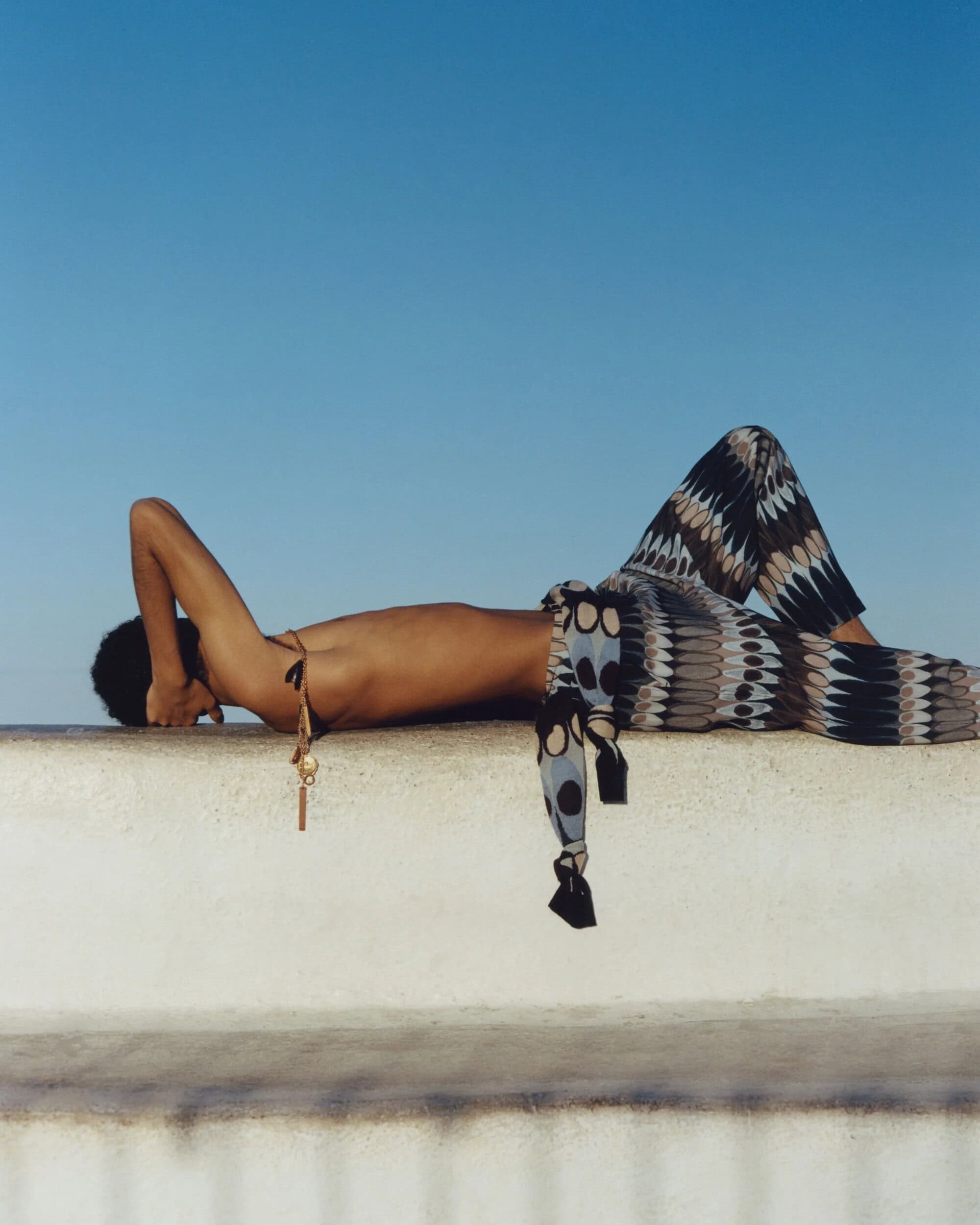 A tanned adult lies on a white wall outdoors under a blue sky, wearing patterned pants, a necklace, and sunglasses, in natural lighting.