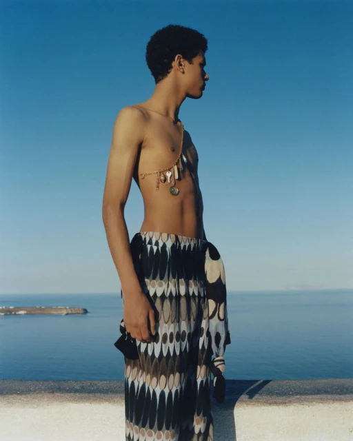 Young adult man poses outdoors on a sunny day with bright blue skies in patterned pants and decorative body chain with ocean in the background.