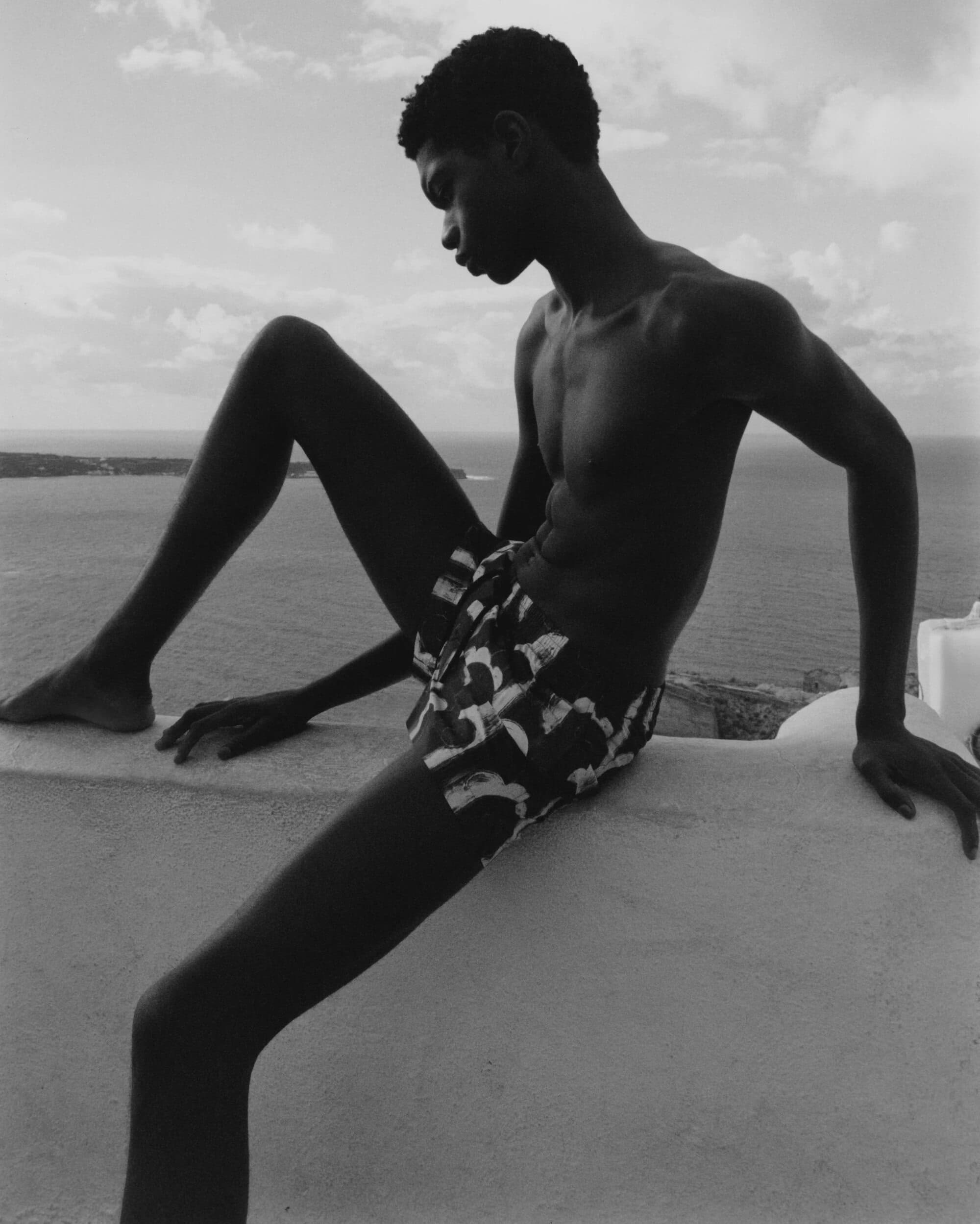Monochrome portrait of a shirtless young Black man in patterned shorts sitting on a white wall with a coastal view in the background.