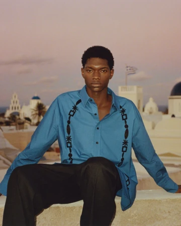 Man in Santorini, Greece poses on a rooftop wearing a blue shirt with black embroidered detail, and black pants with white buildings.