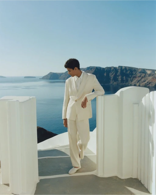 Person in an off-white suit leans against a wall overlooking a blue ocean with a mountainous coastline under a clear sky on a sunny day.