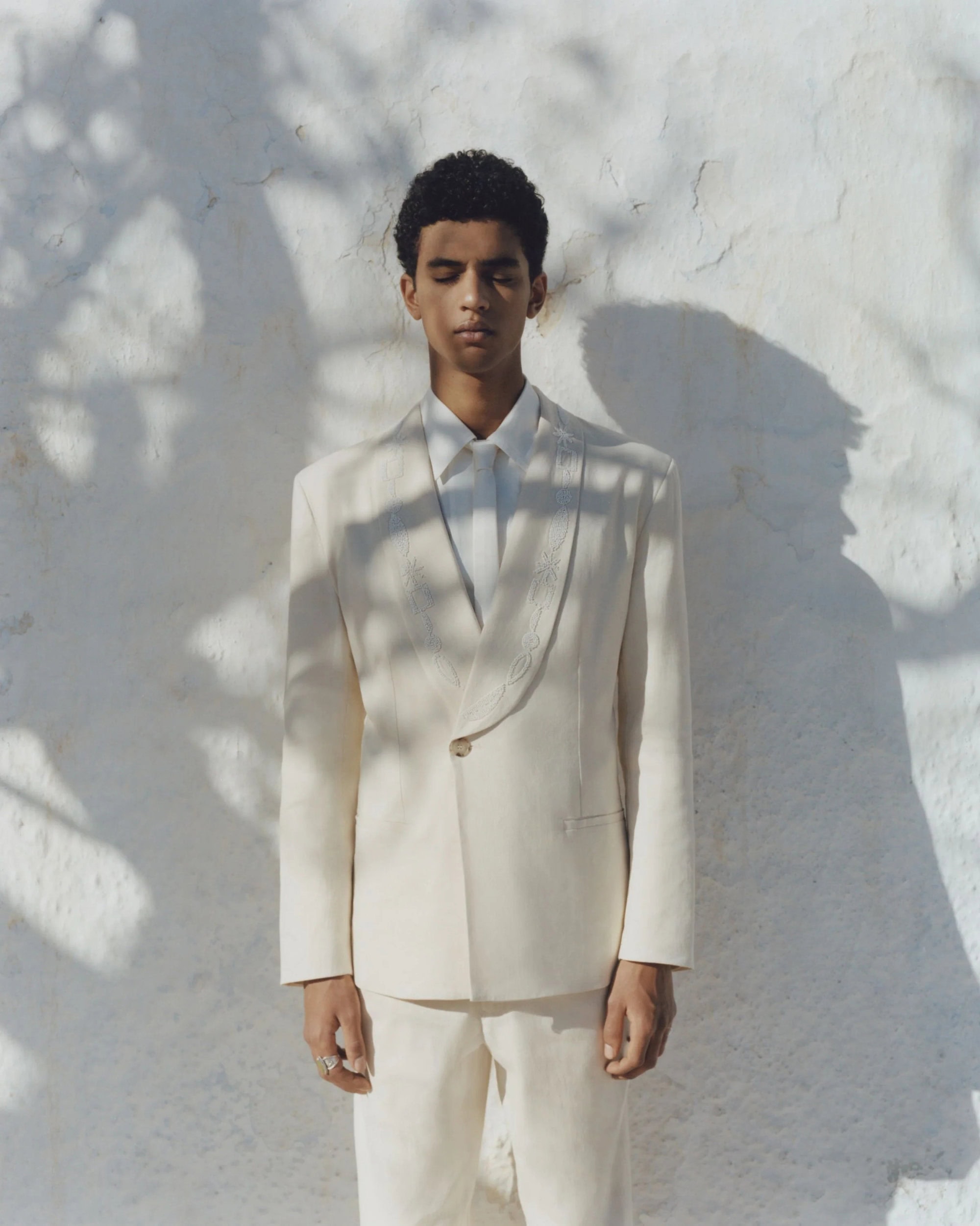 Adult male fashion model in tailored ivory suit posing against a sun-dappled wall in outdoor setting with shadows. Neutral color palette.