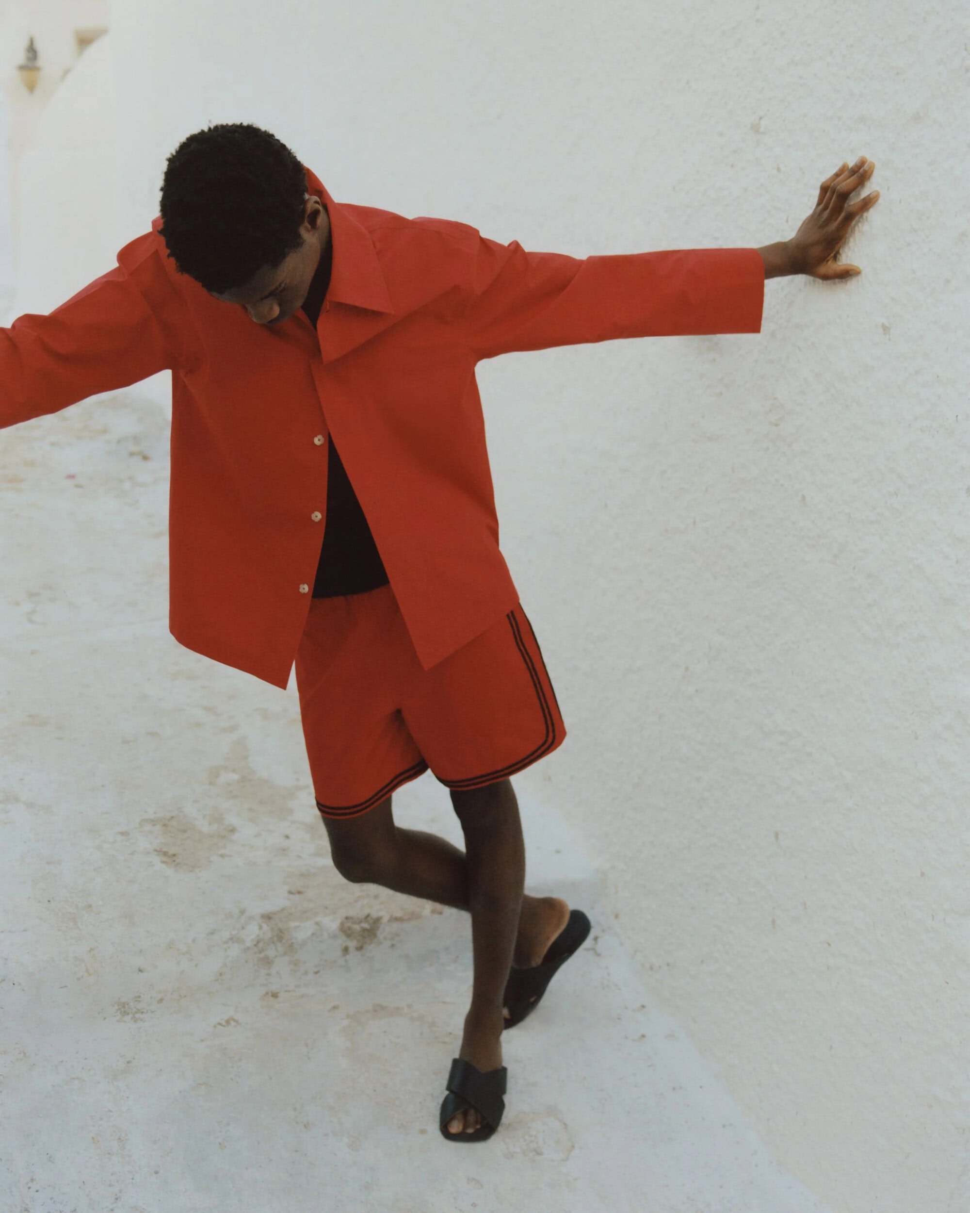 Man in red shirt and shorts leans against a white wall on a bright day, showcasing relaxed fashion and casual style.