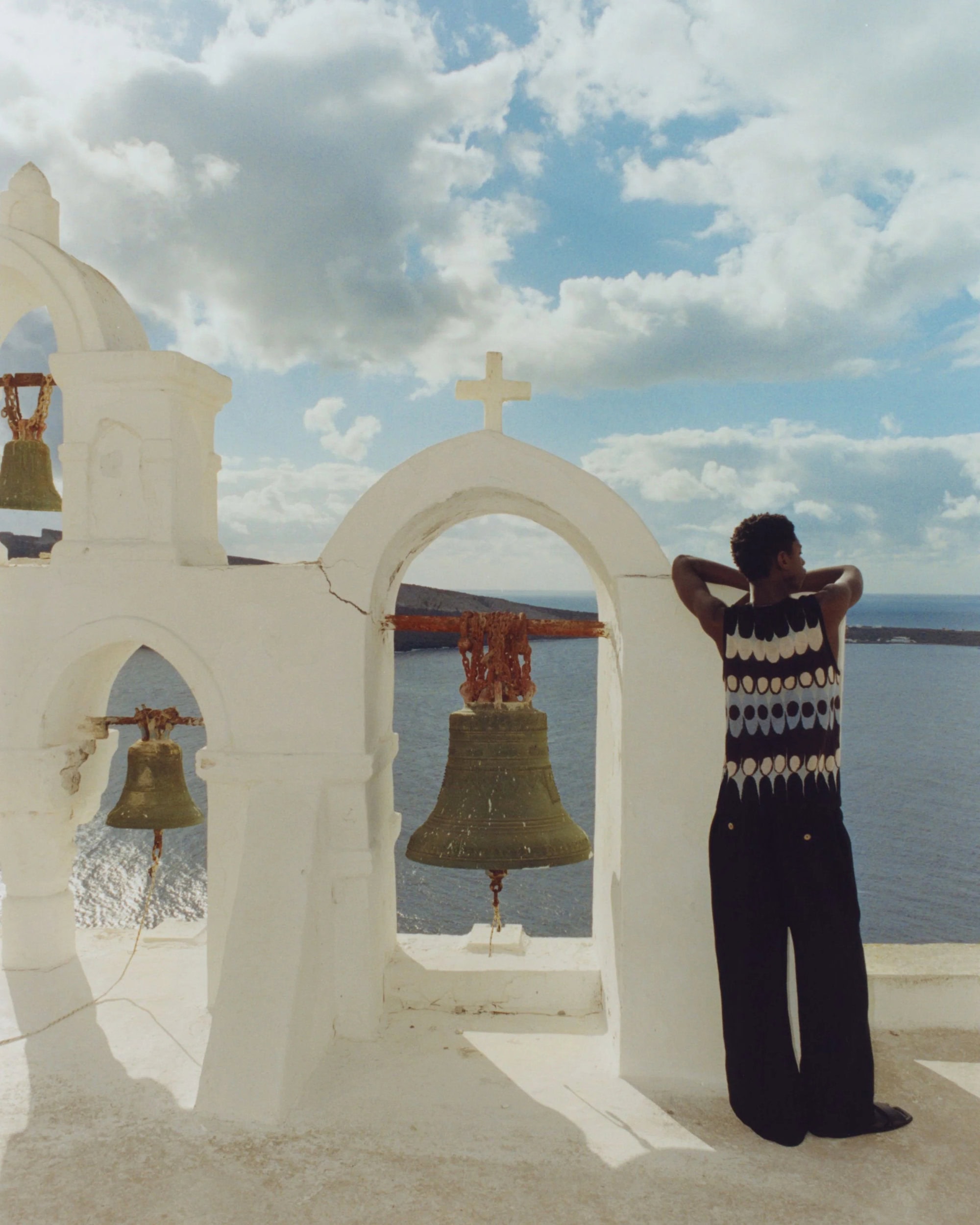 Adult in stylish top stands near whitewashed church bell tower with sea view, under a partly cloudy blue sky in daylight.